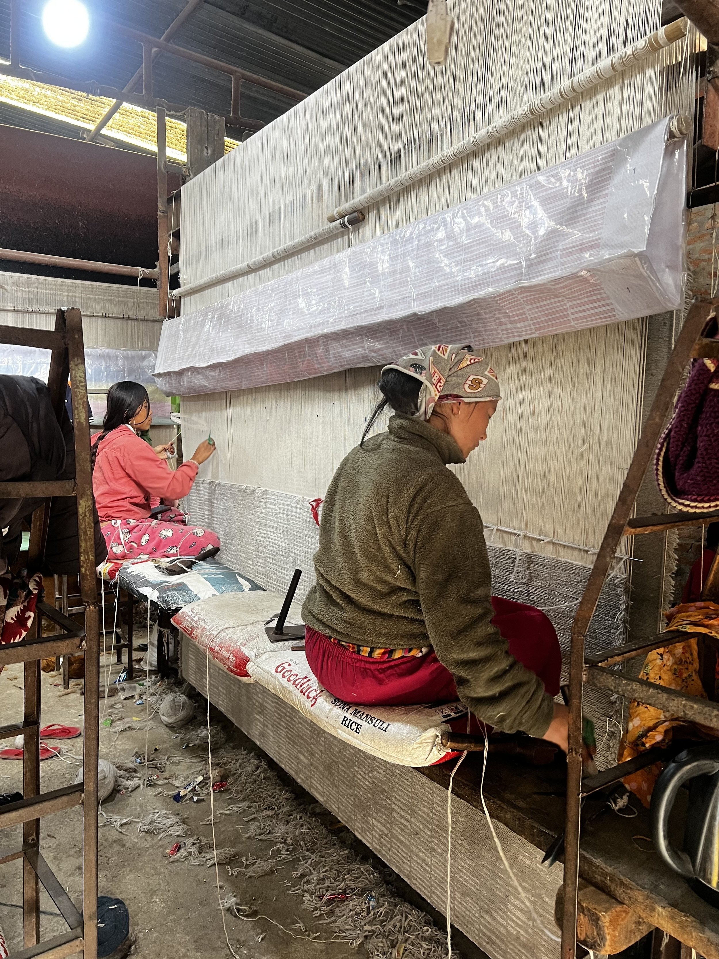Two women working on a loom in a workshop, one sitting on a bench and the other on a cushion, surrounded by various tools and materials.
