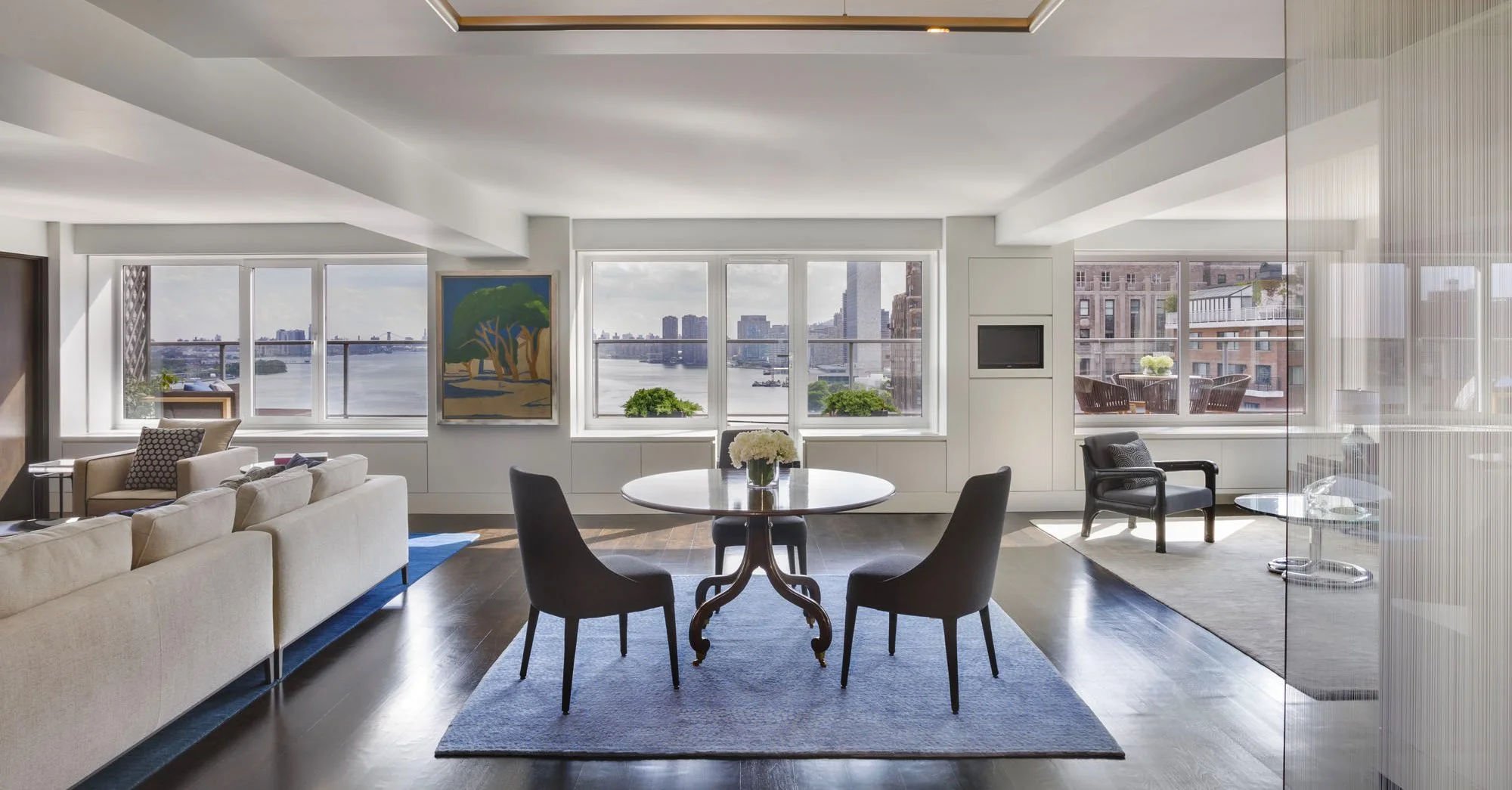 Living room with large windows overlooking a river view, beige sofa, black dining chairs around a round table with white flowers, artwork on the wall, and a balcony with outdoor seating.