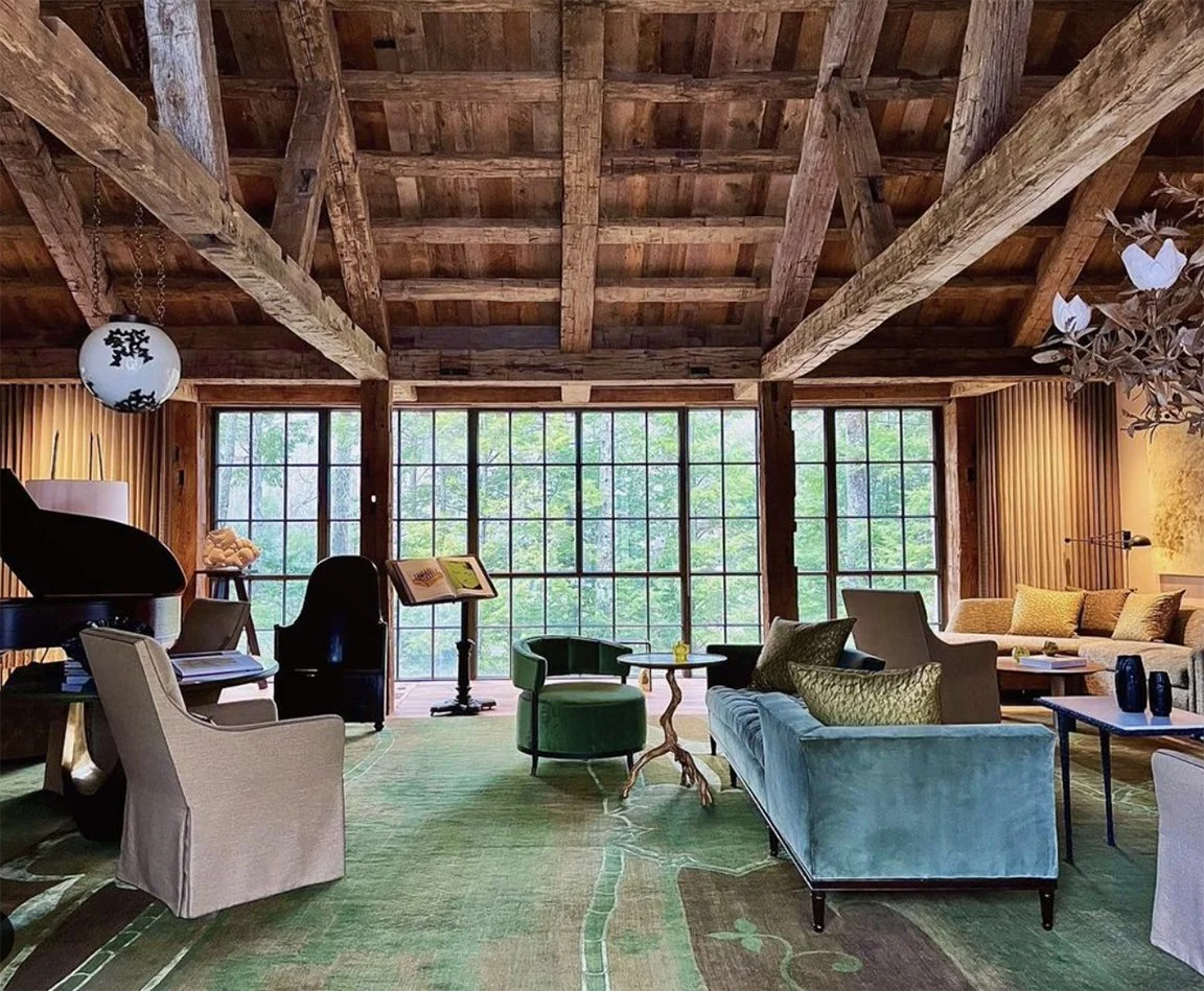 Living room with wooden ceiling, large windows, and a mix of modern and vintage furniture, including a teal sofa, green armchair, beige chairs, and a black grand piano.