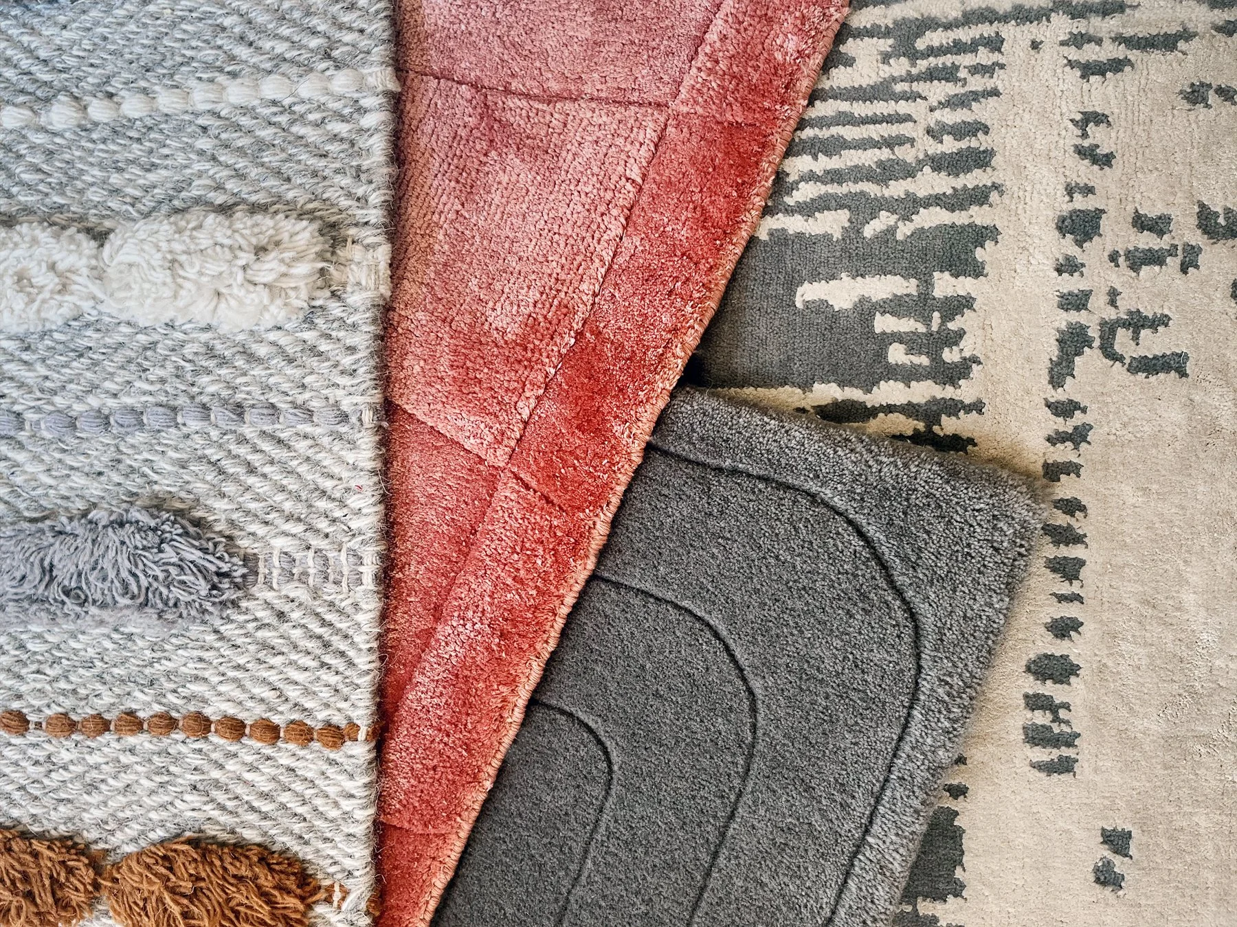 Various folded rugs and carpets with different textures and patterns in neutral, gray, red, and beige colors, displayed on a surface.