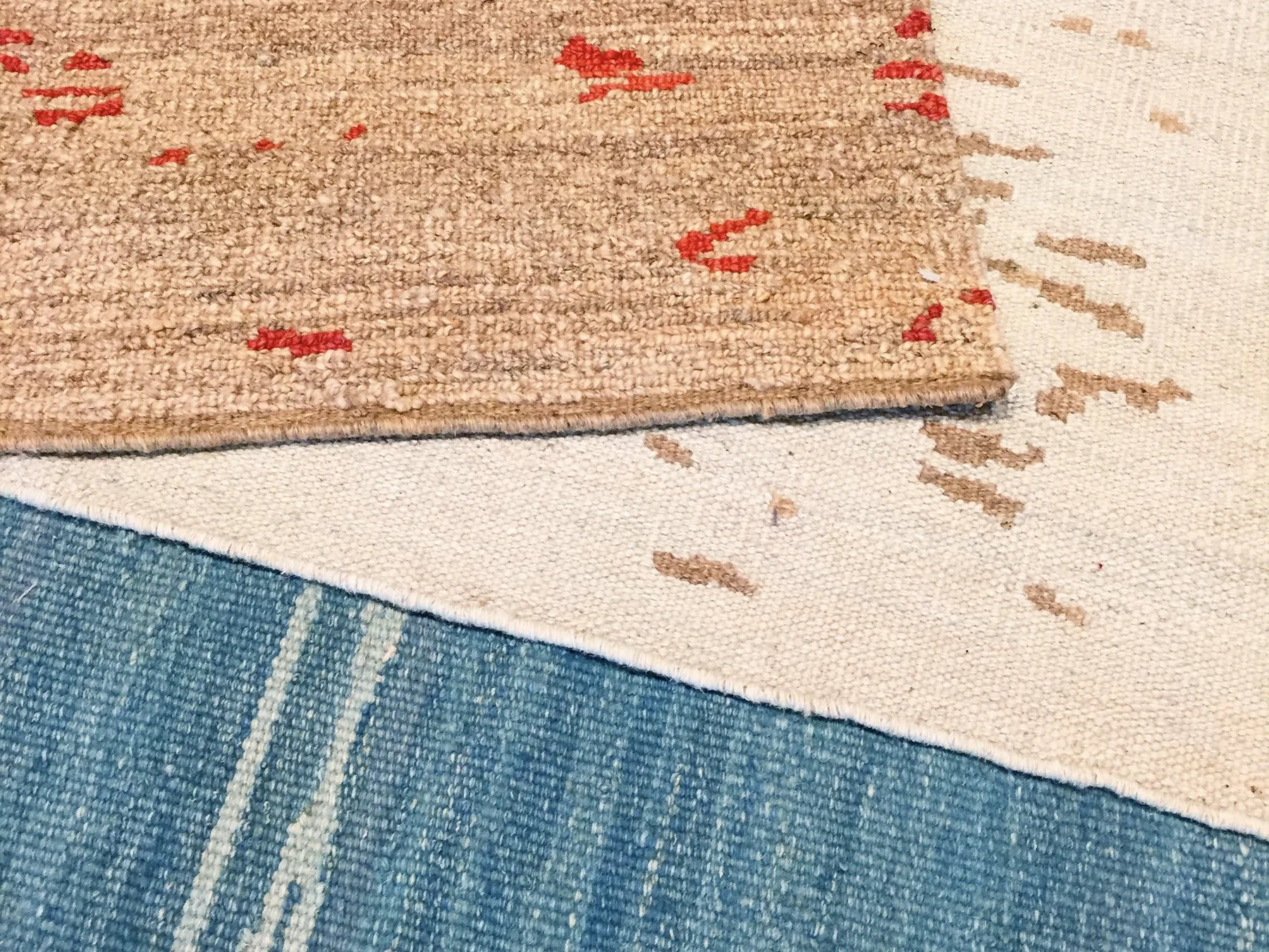 Close-up of a beige flatweave with red and brown accents, partially overlapping a white and brown speckled flatweave on a blue flatweave rug.