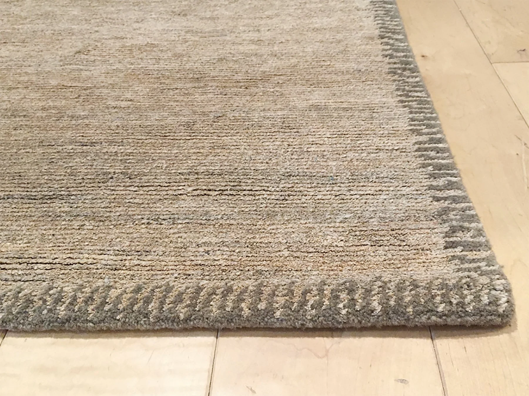 Close-up of the corner of a beige area rug with decorative border on a light wooden floor.