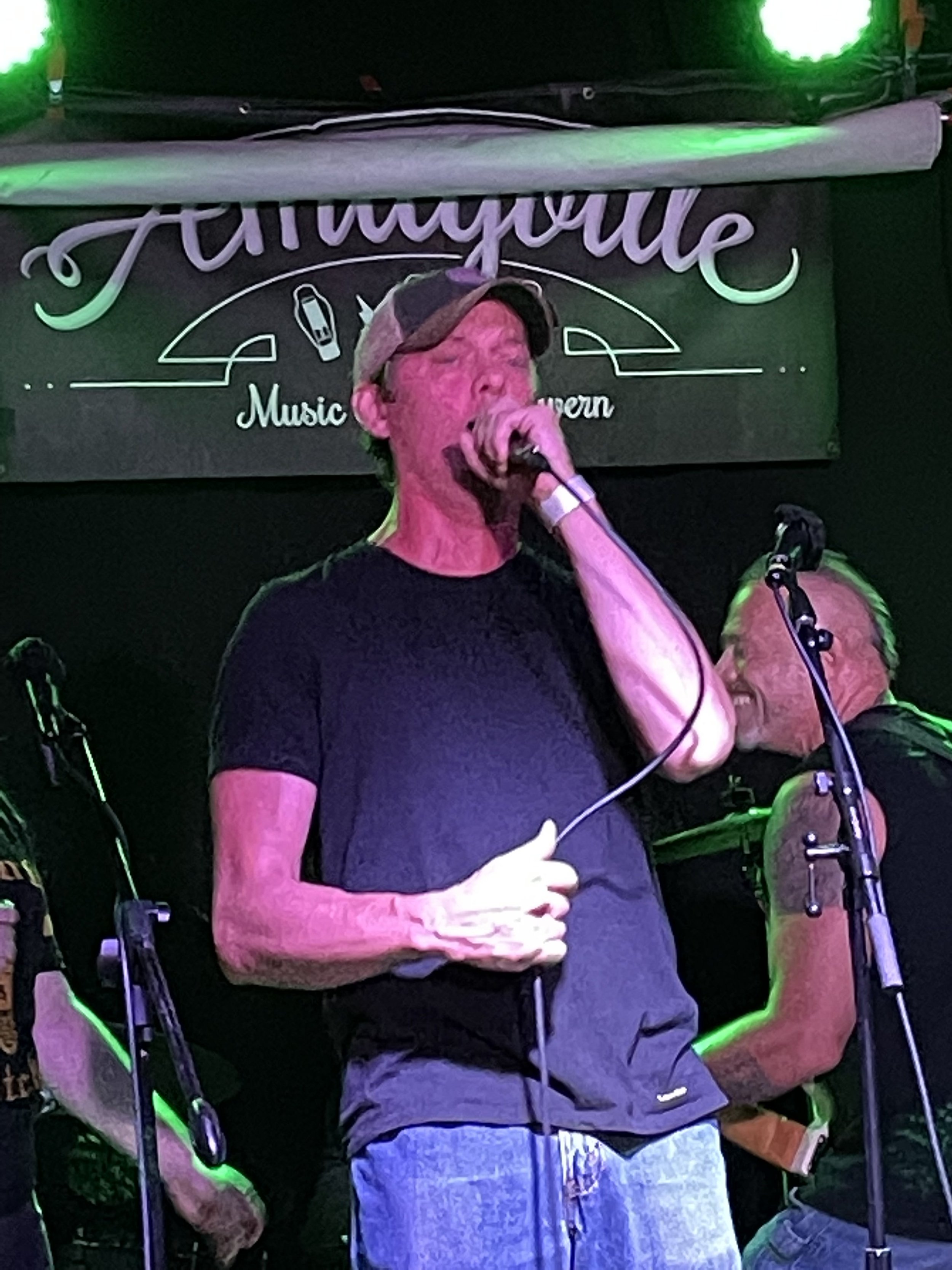 A person singing into a microphone on stage at a venue called Amityville Music Hall. The person is wearing a cap and a black shirt. There are other band members in the background with equipment like microphones and instruments.
