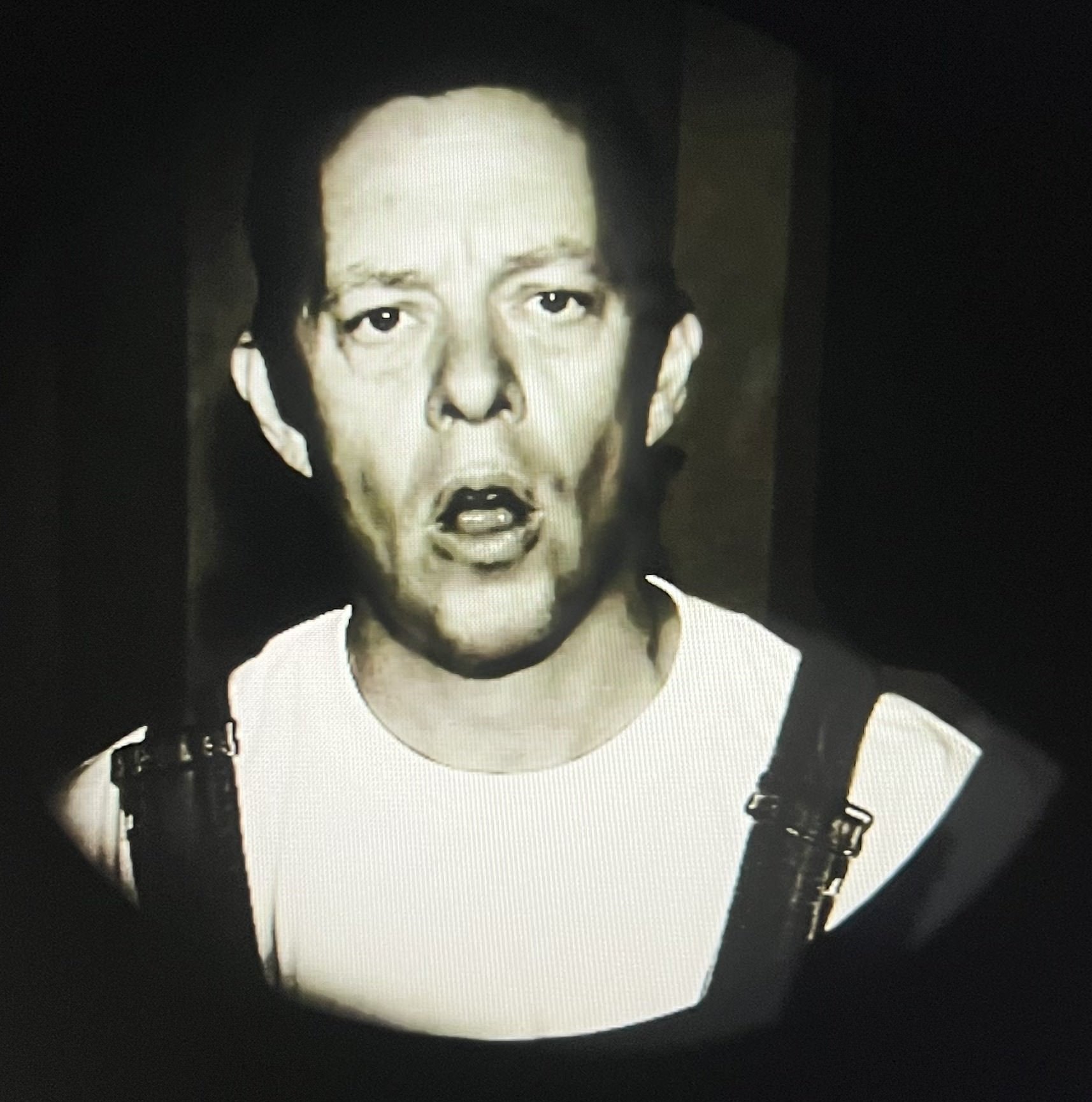 Black and white image of a person wearing overalls and a white shirt, with an open mouth expression, looking directly at the camera.