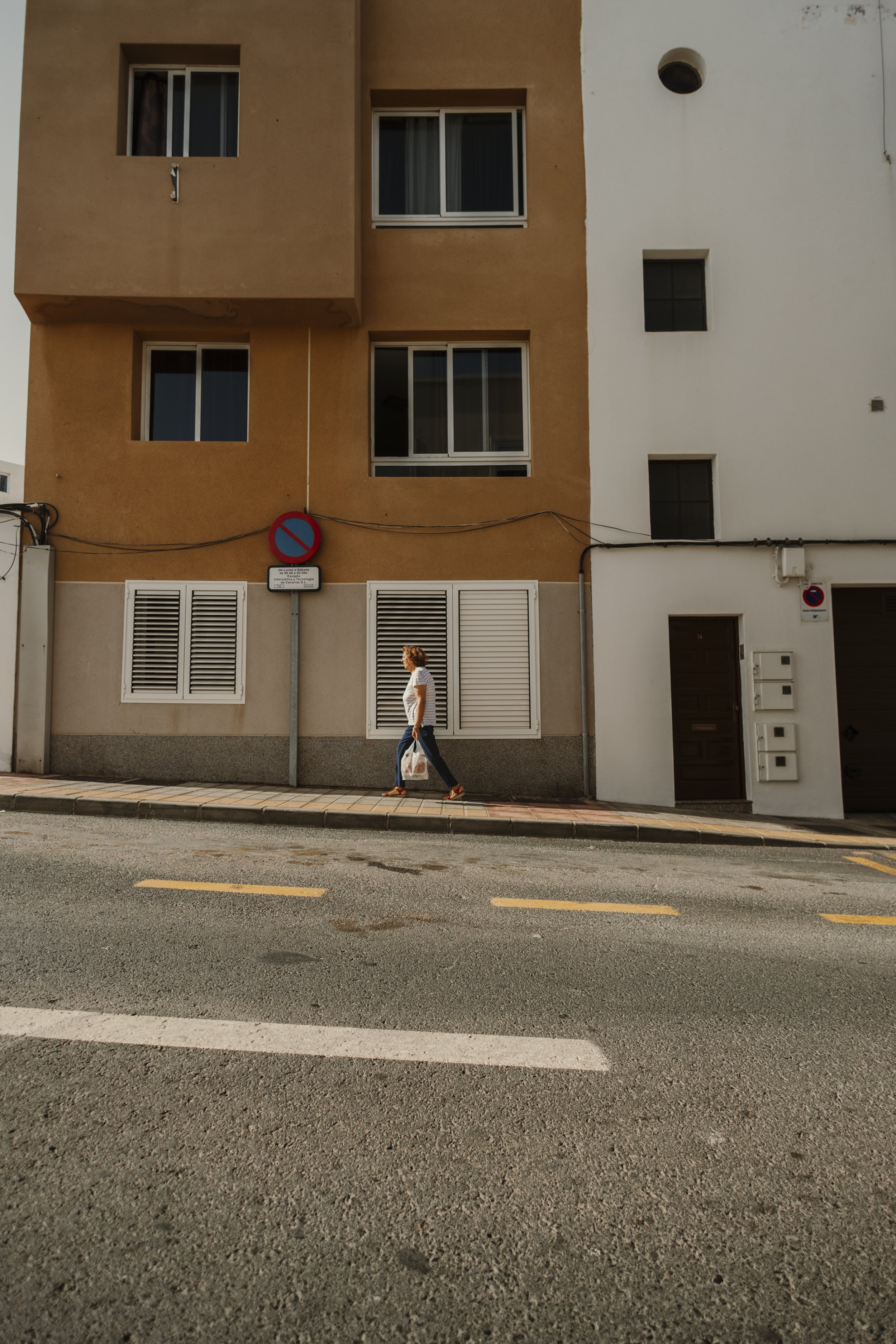 Una donna cammina sul marciapiede davanti a un edificio residenziale con facciata beige e bianca, con targhetta di divieto di sosta