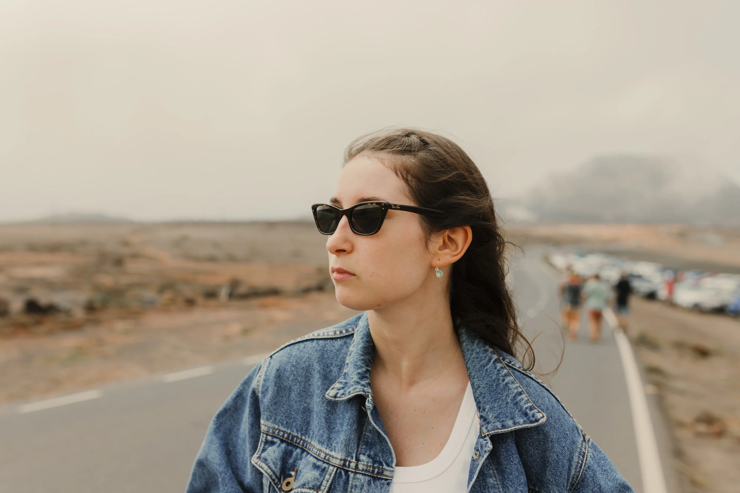 Ragazza con occhiali da sole e giacca di jeans in un paesaggio arido e aperto, con persone e auto sullo sfondo.