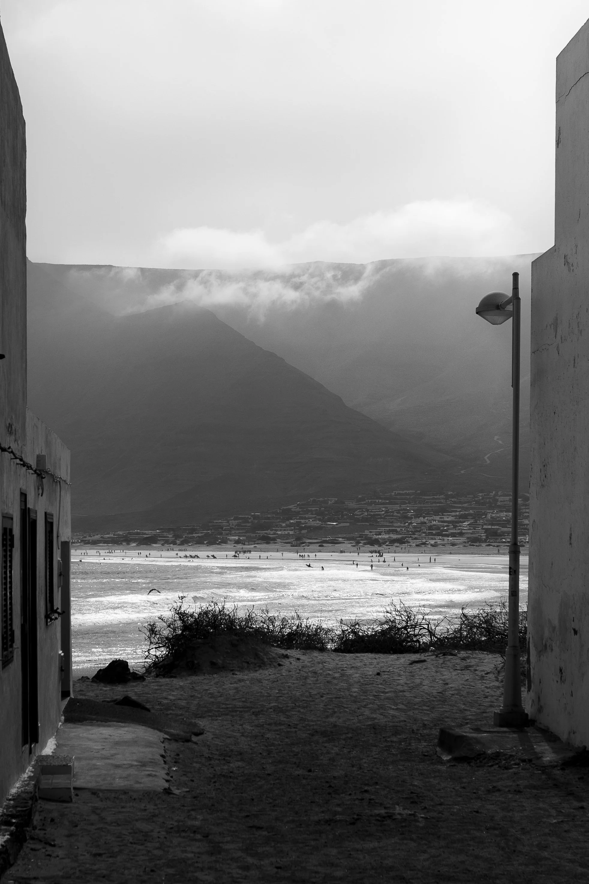 Veduta di una spiaggia con montagne sullo sfondo, visualizzata attraverso un'apertura tra edifici, in bianco e nero.