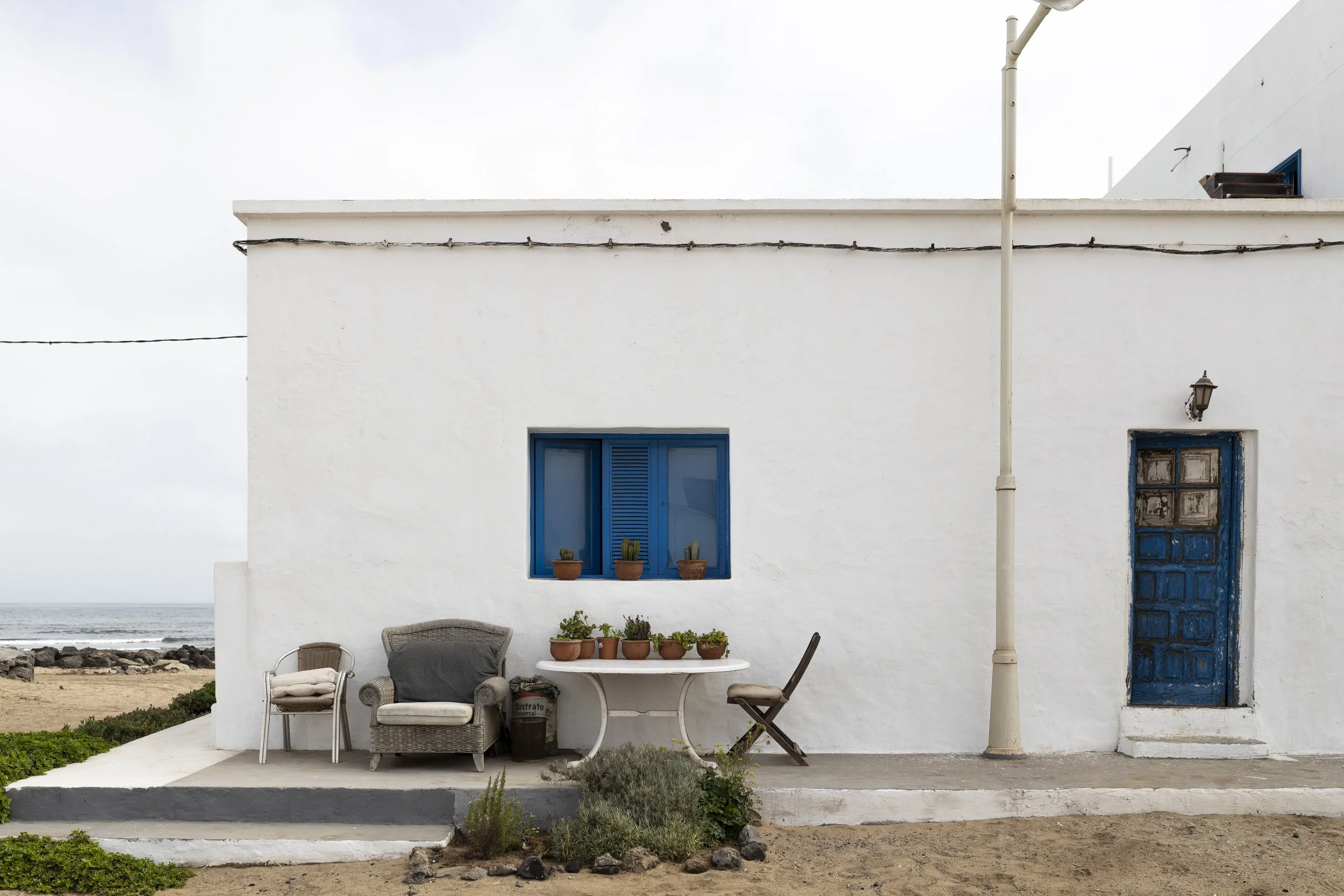 Casa bianca con porta e finestra blu, arredamento esterno con sedie e vaso di piante, vicino al mare.