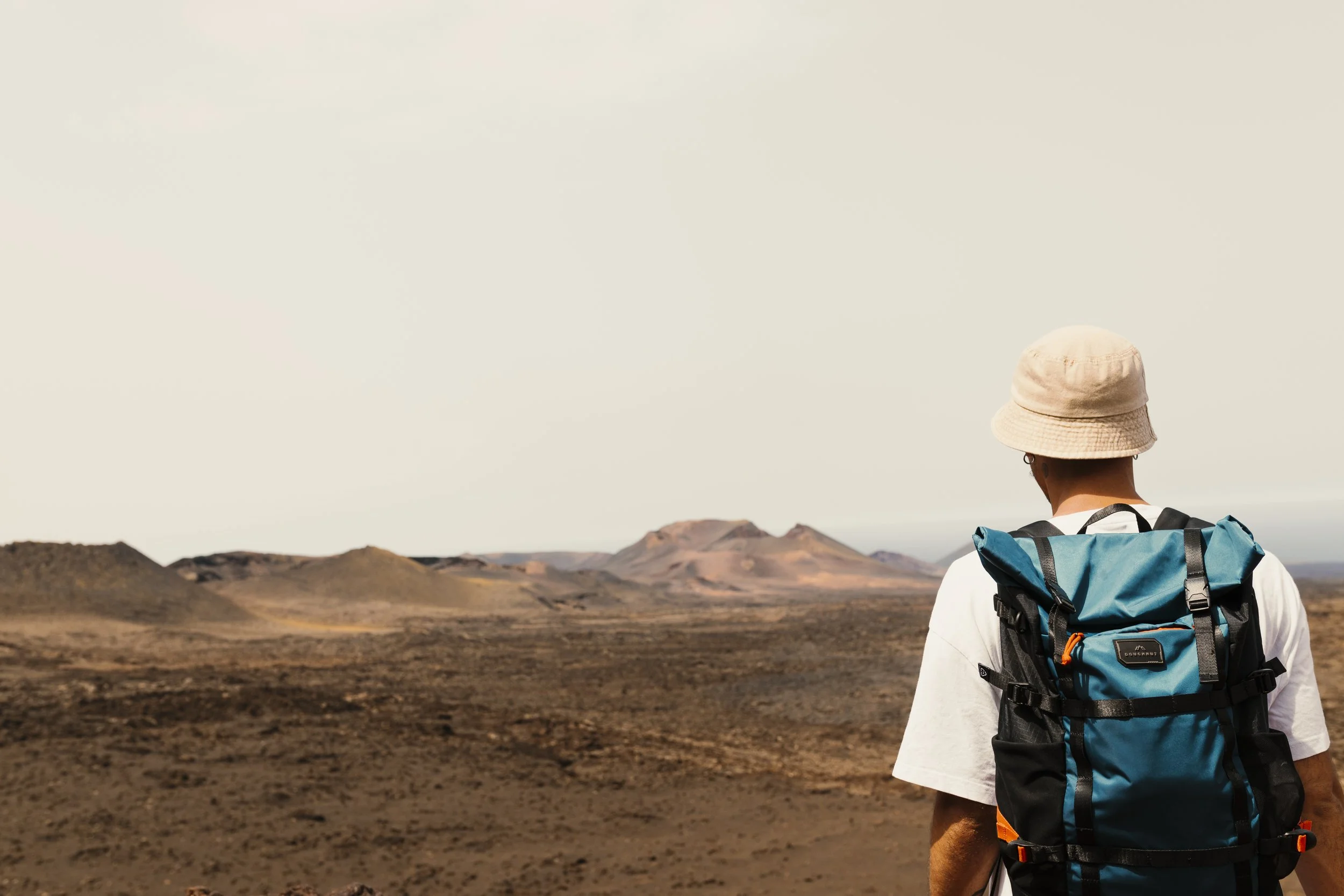 Persona con cappello e zaino blu che guarda un paesaggio desertico con colline e montagne in lontananza.