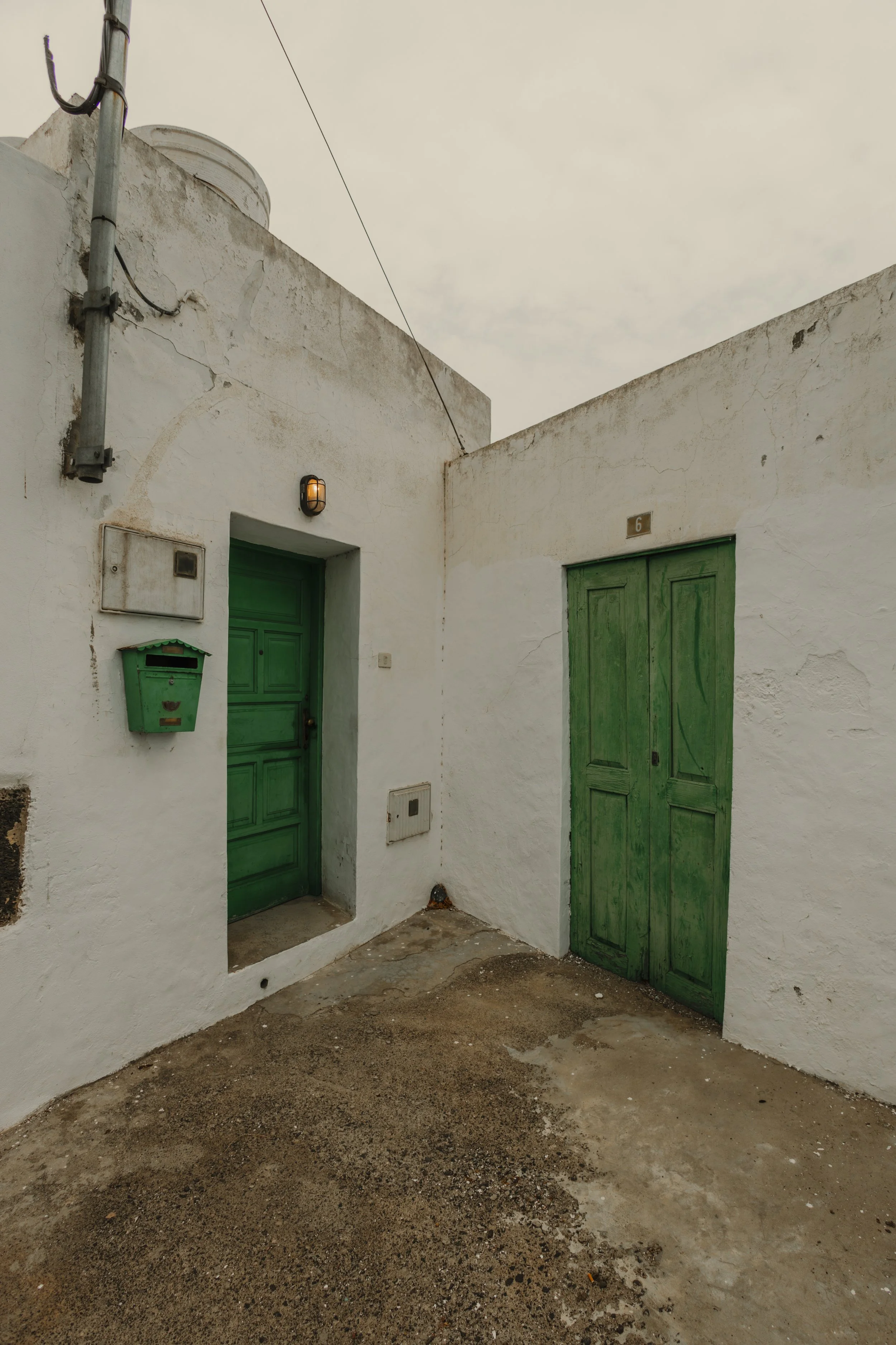 Cortile con muro bianco, due porte verdi, una con finestra sulla parte superiore, e una cassetta delle lettere verde, sotto un lampione acceso, in un ambiente rustico con pavimentazione usurata.