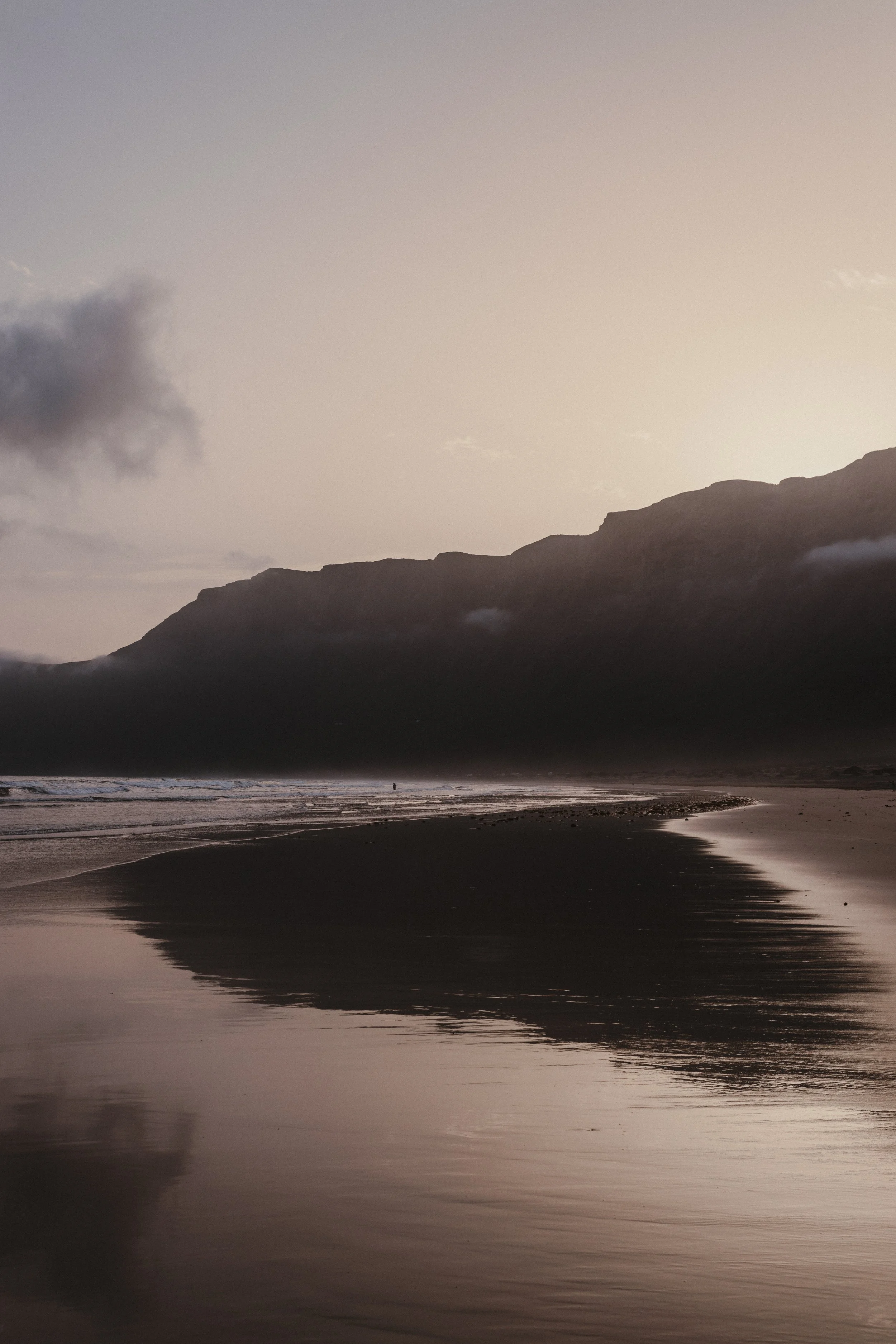 Paesaggio al tramonto con montagna, spiaggia e mare calmo che riflette il cielo rosa.