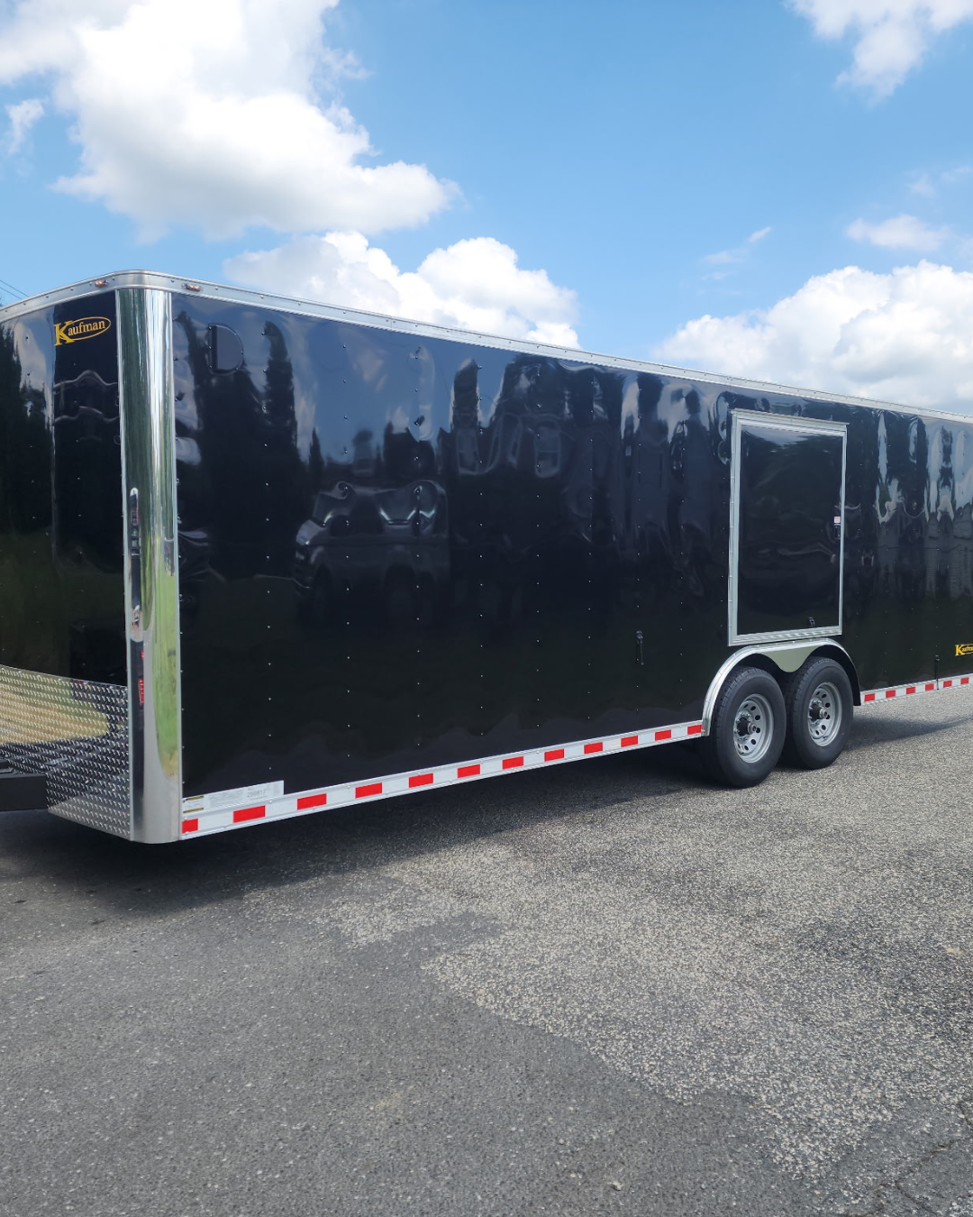A large black enclosed trailer with dual axles parked on a paved surface under a partly cloudy sky.