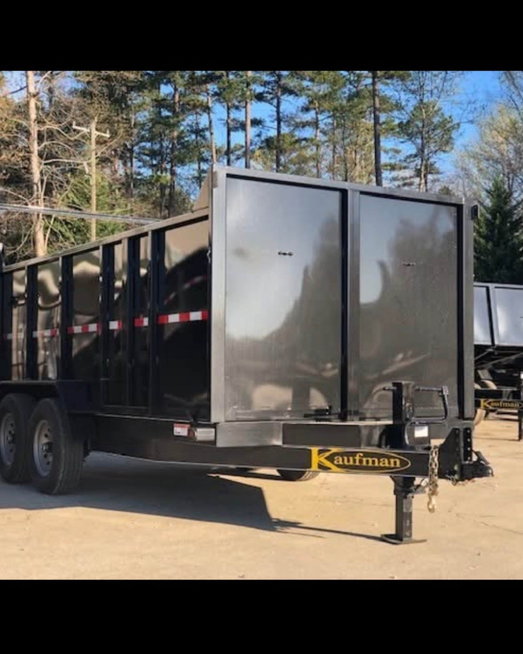 A black Kaufman trailer with a silver metal enclosed back doors, parked on a dirt surface with trees and blue sky in the background.