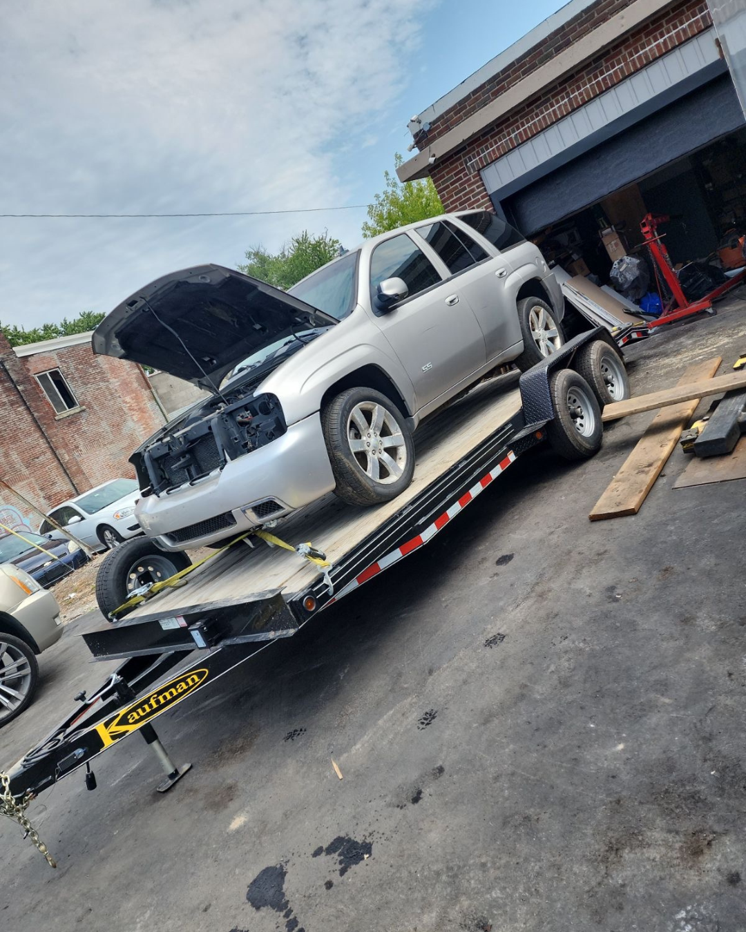 Silver SUV with open hood loaded on a black flatbed trailer in a driveway near a garage, with car repair equipment and wood planks around.