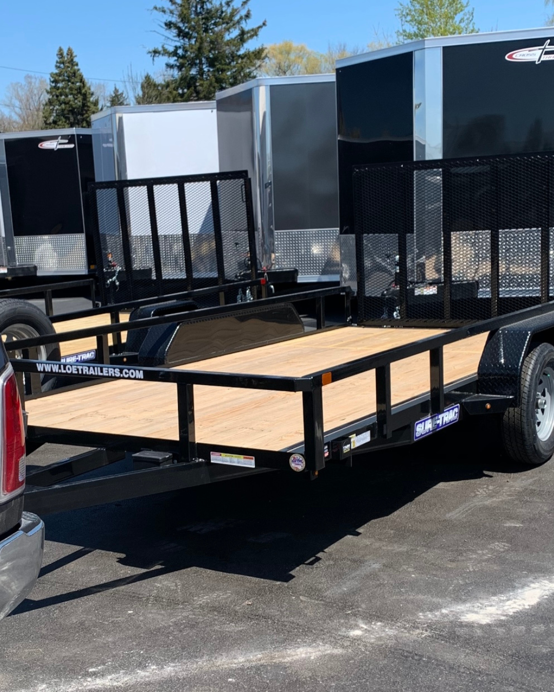 Black flatbed trailer with wood deck parked on asphalt, with black metal side rails and black mesh ramps, attached to a pickup truck. Several enclosed trailers are visible in the background, under a clear blue sky with some trees.
