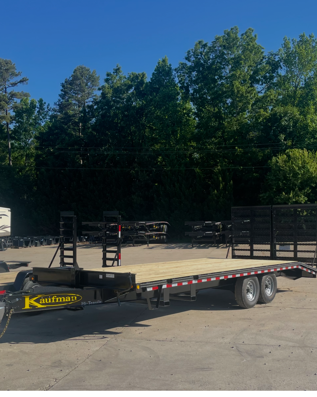 A flatbed trailer parked on a concrete surface with trees and blue sky in the background.
