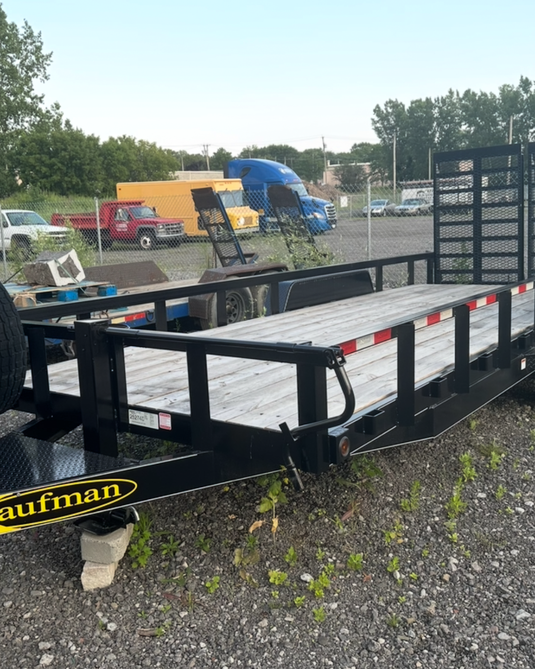 A flatbed trailer with a black metal frame and wooden deck, parked on gravel. In the background, there are several vehicles including a yellow truck with a blue cab, a red truck, and some cars, surrounded by a chain-link fence.