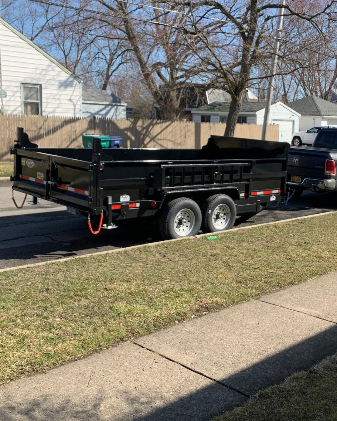 A black utility trailer attached to a black pickup truck parked on a residential street. The trailer has double wheels, red safety chains, and is empty.