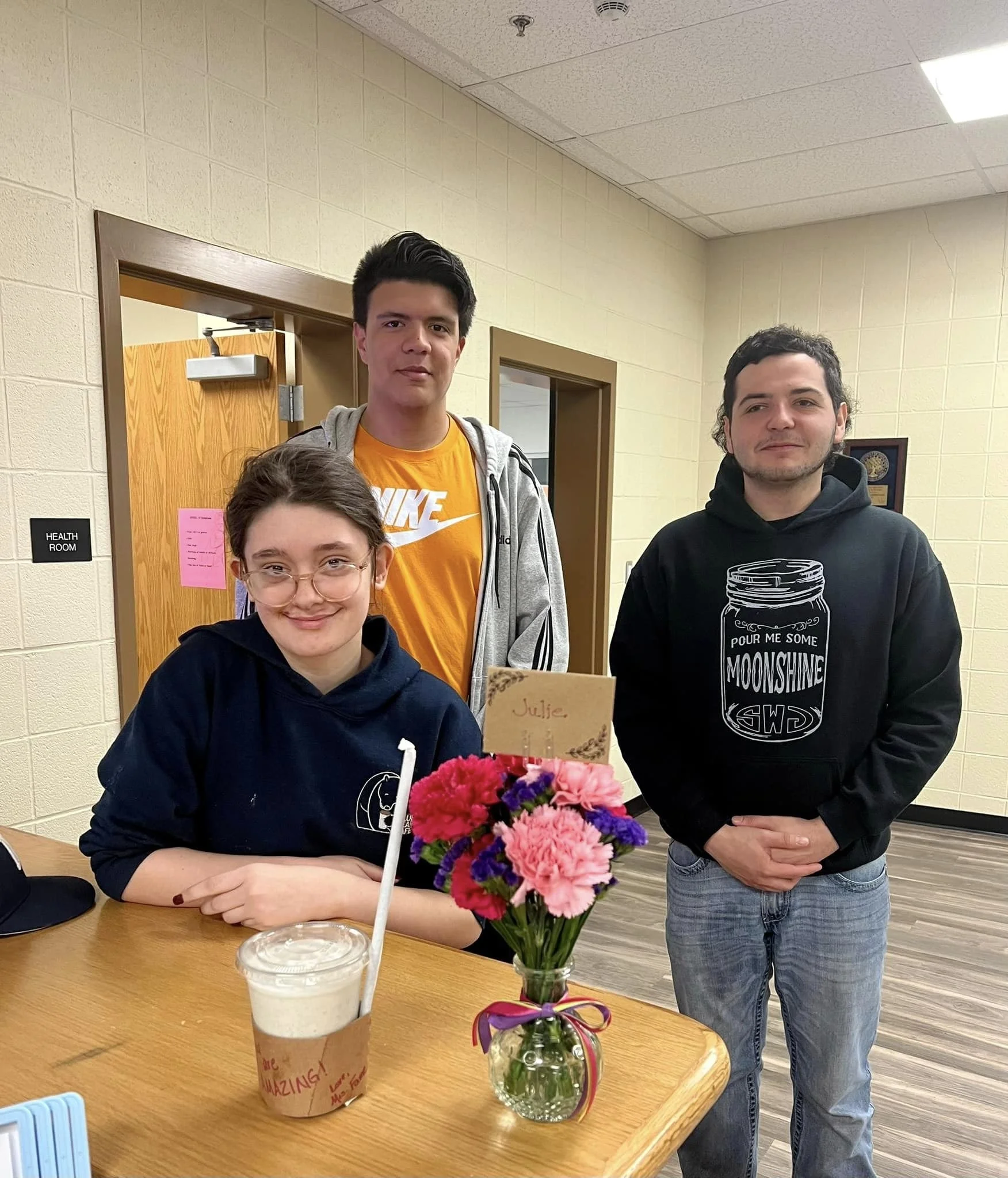 Three people in a room, one sitting at a table with a drink and a flower arrangement, two standing behind them. The room has wooden doors and a sign labeled 'Health Room.'