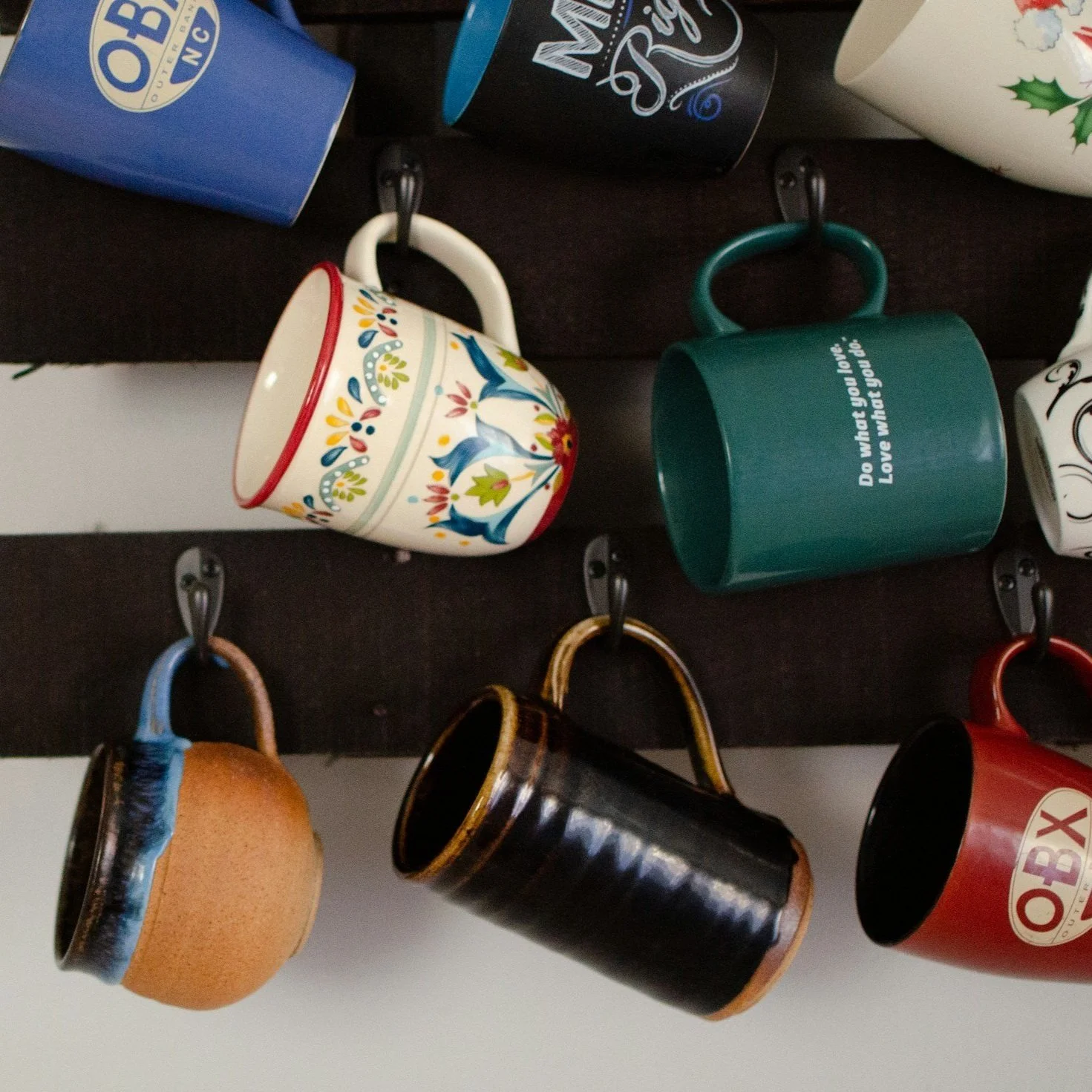 Various mugs hanging on hooks, including a blue 'OBX' mug, a floral patterned mug, a green mug with a white inscription, and a brown ceramic mug.