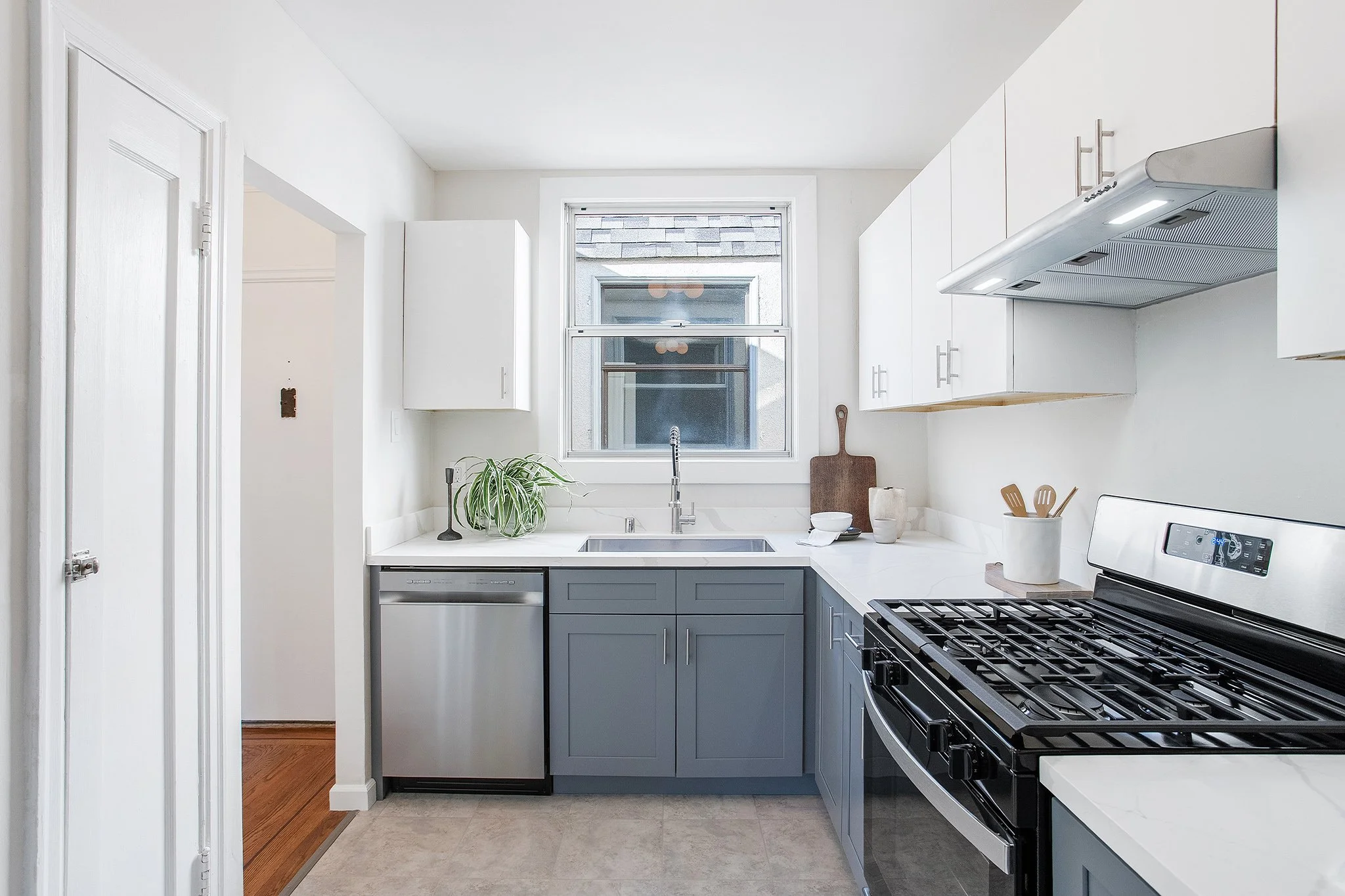 Modern kitchen with white upper cabinets, gray lower cabinets, a stainless steel dishwasher, black stove, and a window above the sink. There are kitchen utensils and decorative items on the countertop.
