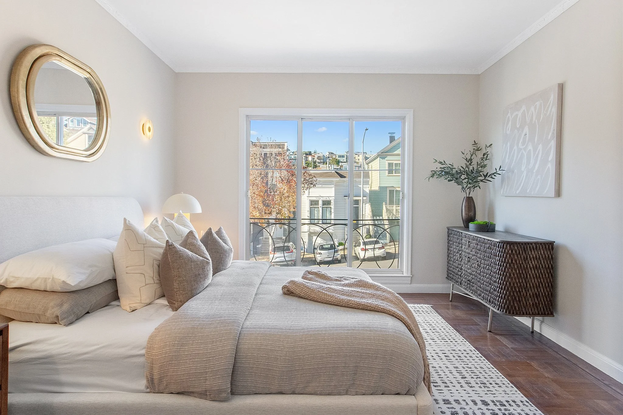 Bright bedroom with a large bed, decorative pillows, a nightstand with a lamp, and a sliding glass door leading to a balcony with parked cars and neighboring houses visible outside.