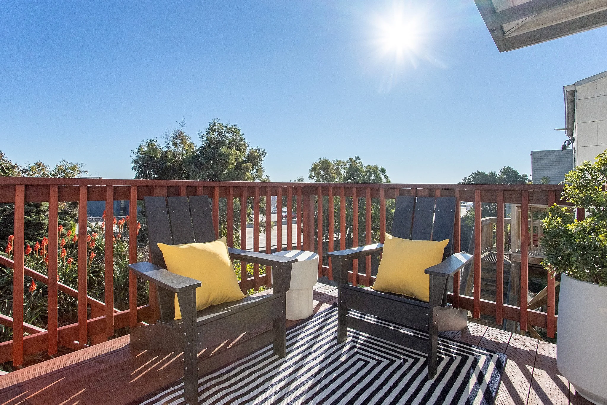 A sunlit outdoor balcony with two black wooden chairs each with a yellow pillow, a striped black and white rug, and a large white planter with a green plant, enclosed by a brown wooden railing, with trees and a blue sky in the background.