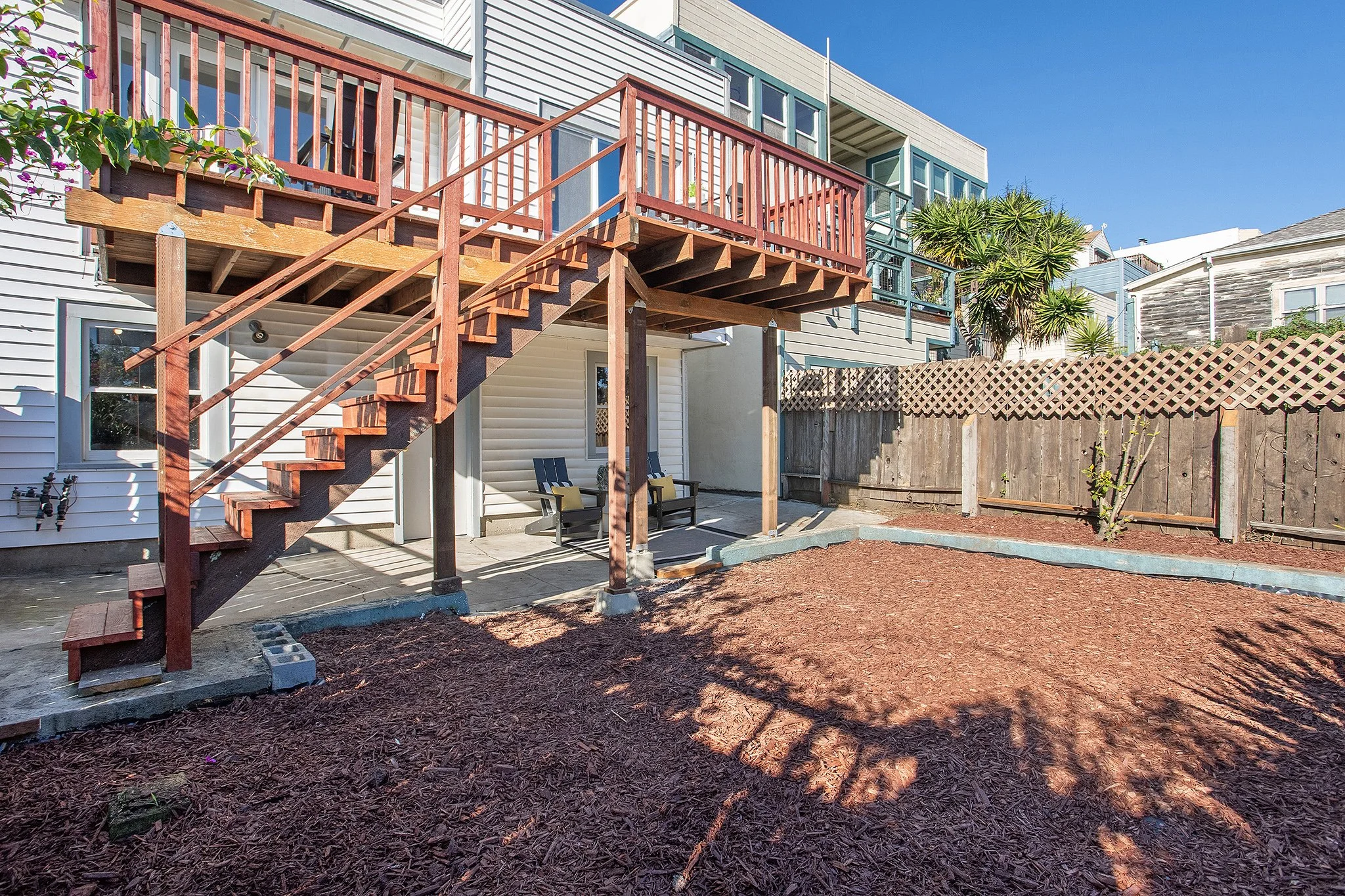 Backyard with a wooden elevated deck with stairs, two chairs on the patio, and a fenced area with some small plants, under a clear blue sky.
