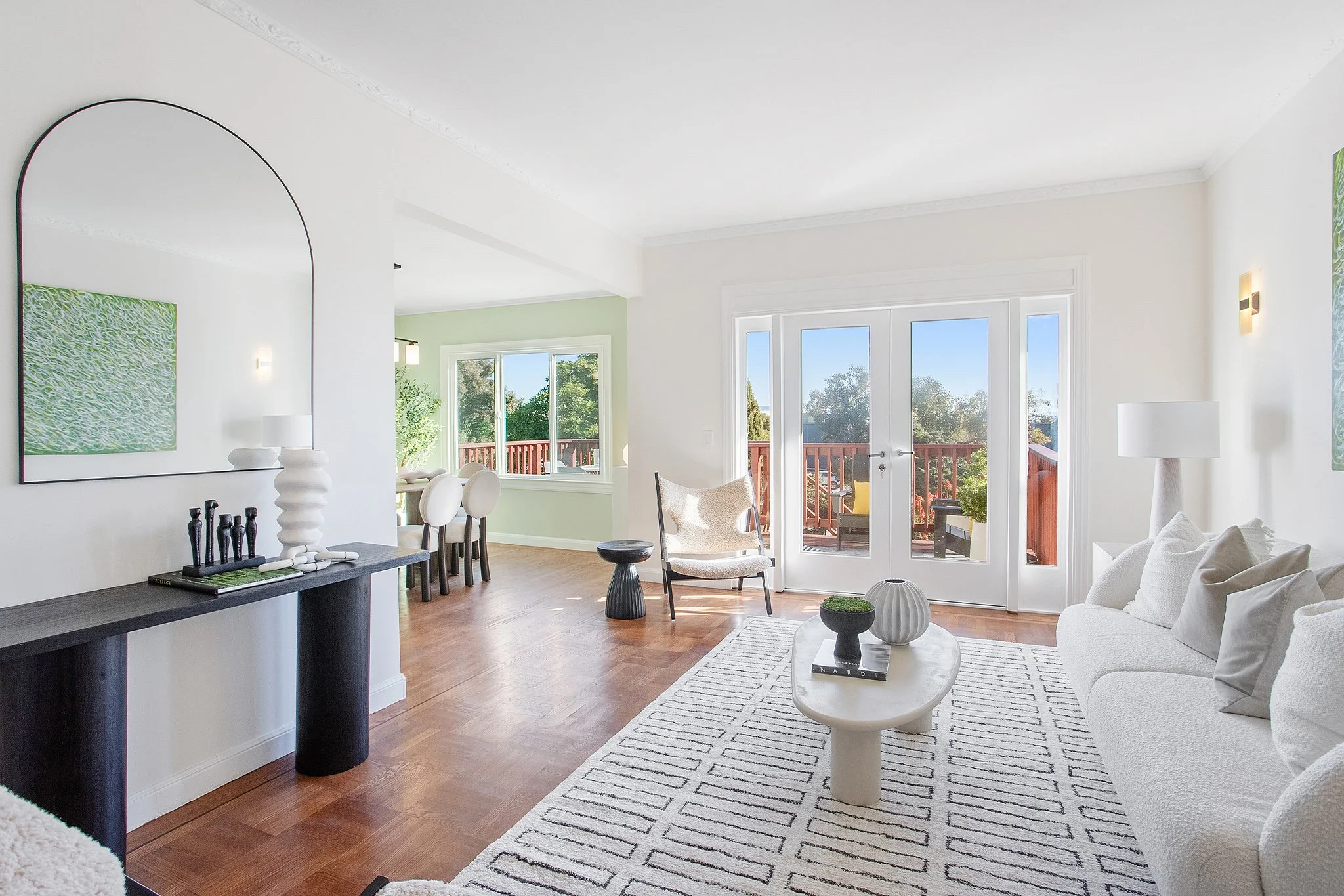 Bright living room with white sofa, round ottoman, coffee table, and large glass doors leading to balcony with outdoor seating. Adjacent dining area with green accent wall and windows.