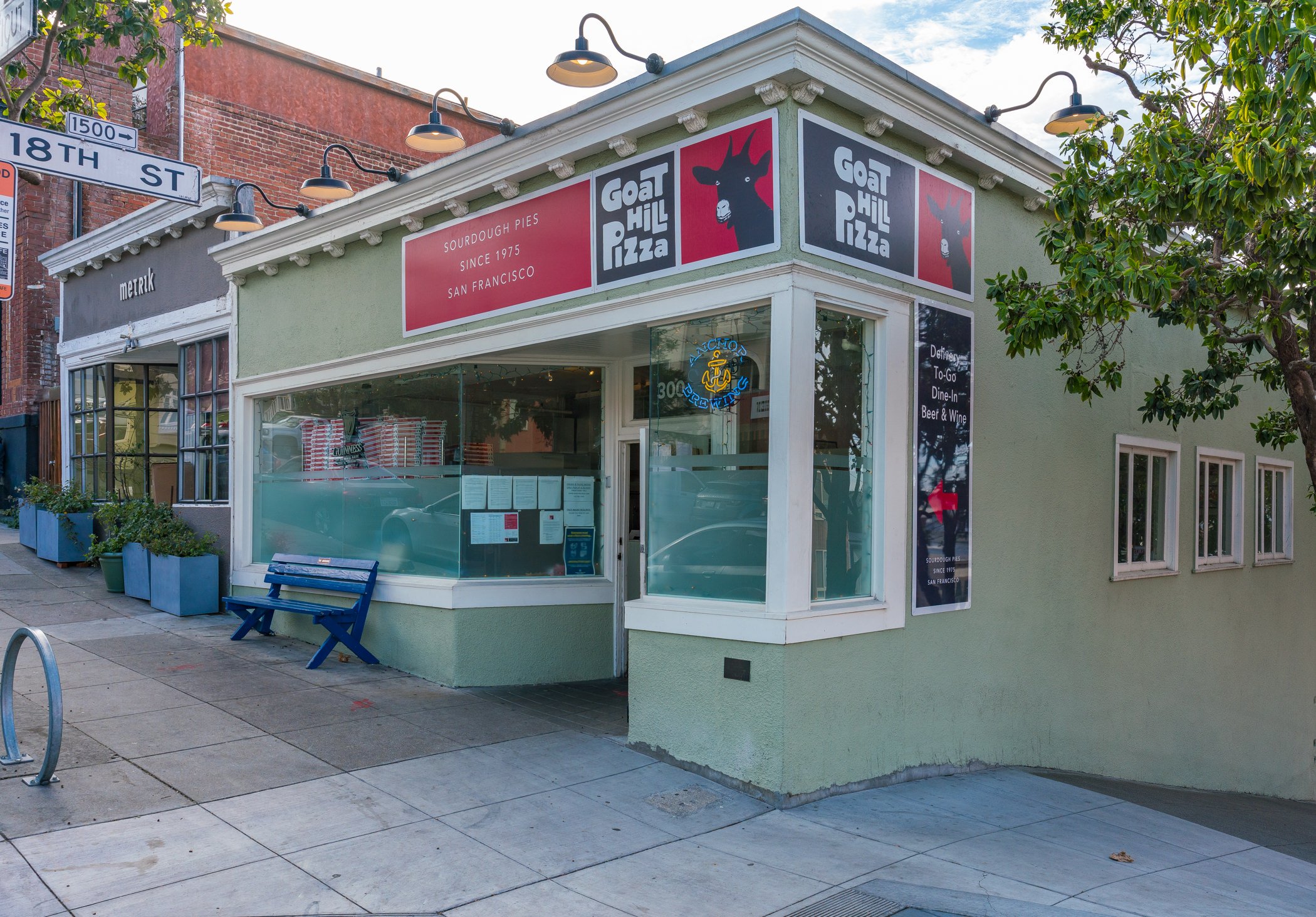 Exterior of a pizzeria called Goar Hill Pizza with signs indicating sourdough pies since 1975 in San Francisco; features large glass windows, a blue bench outside, and a neon sign inside.