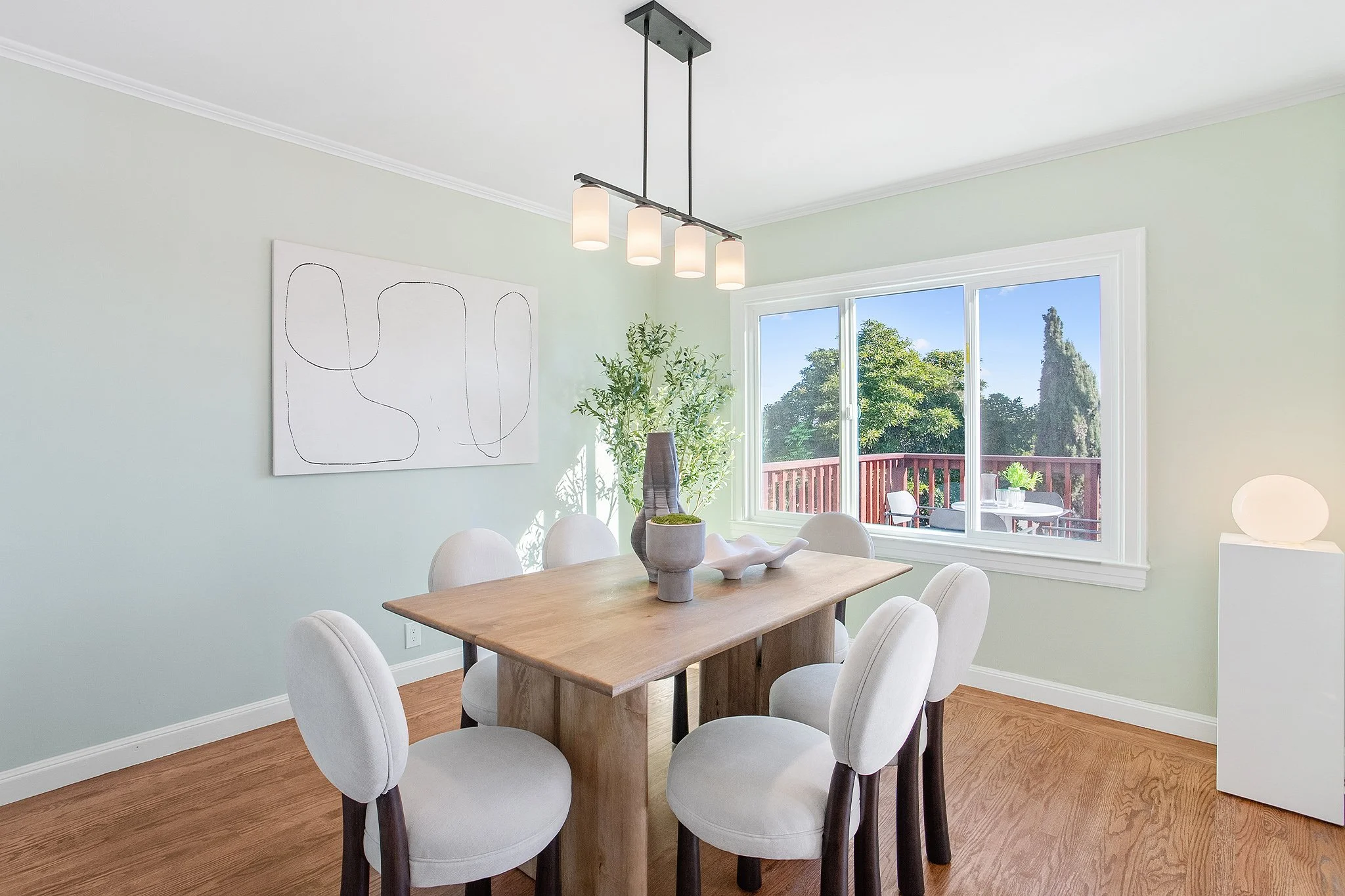 Bright dining room with six white upholstered chairs around a wooden table, decorative vases and sculpture on the table, a large window with an outdoor view, a modern light fixture, and minimal artwork on light-colored walls.