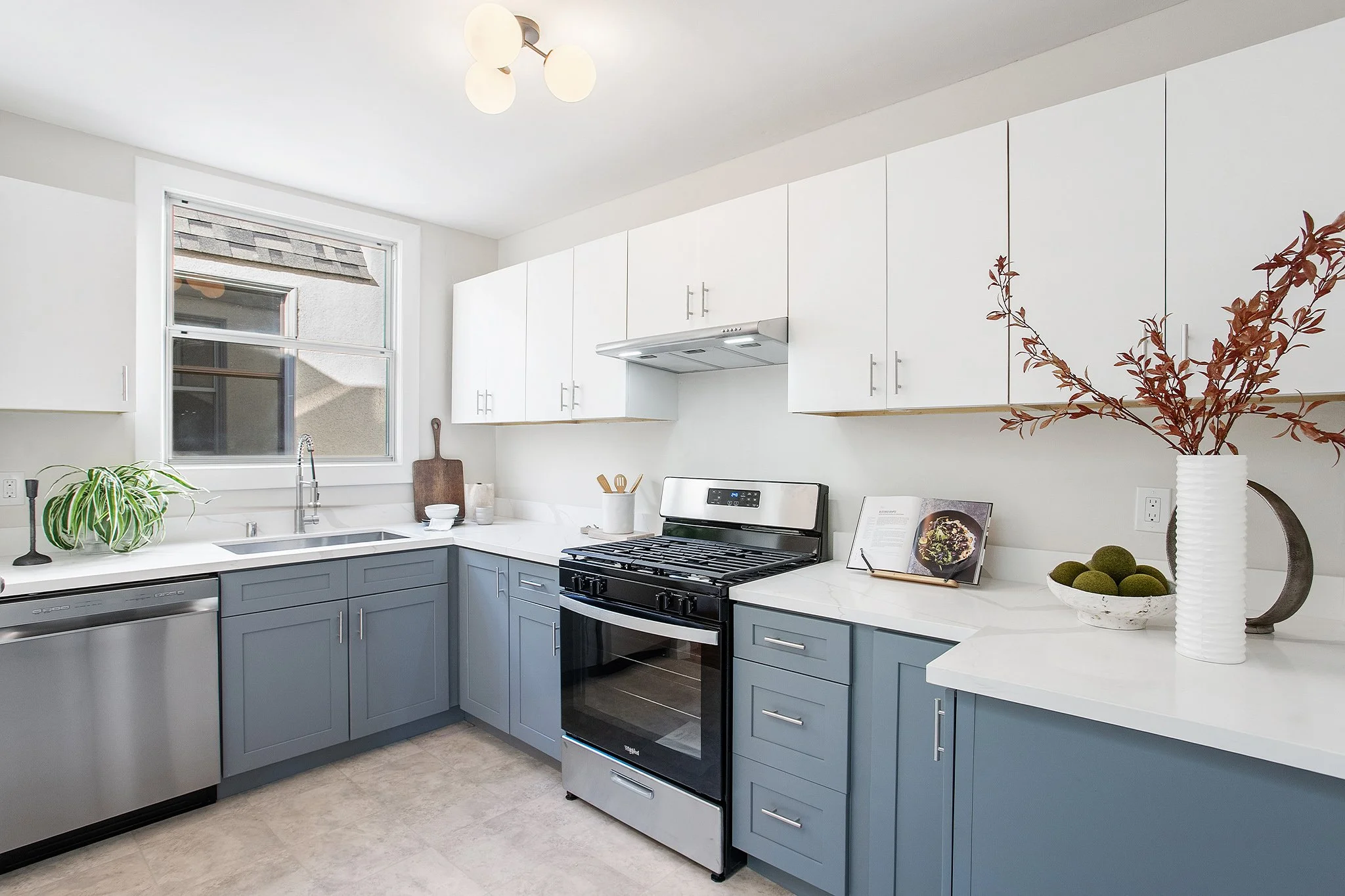 Modern kitchen with white upper cabinets, blue lower cabinets, stainless steel dishwasher and stove, a window above the sink, and decorative items including a large white vase with brown leaves, a small bowl of green moss balls, and an open cookbook.