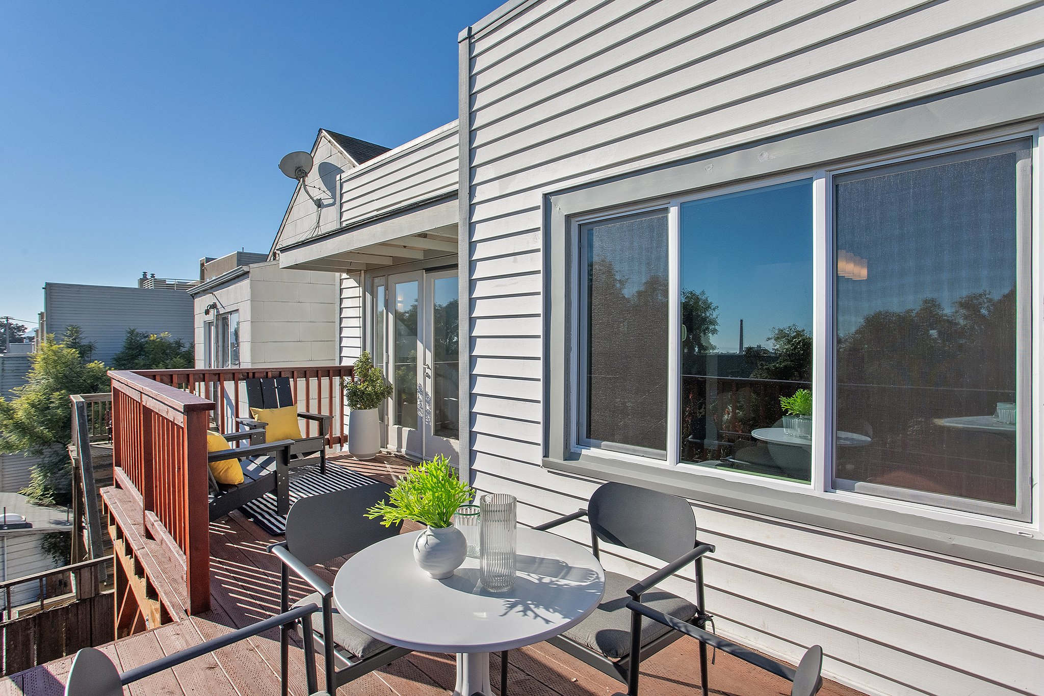 Balcony with outdoor furniture, including a round table with two chairs, a black bench with yellow pillows, and potted plants outside a white house with sliding glass doors and windows. The sky is clear and sunny.