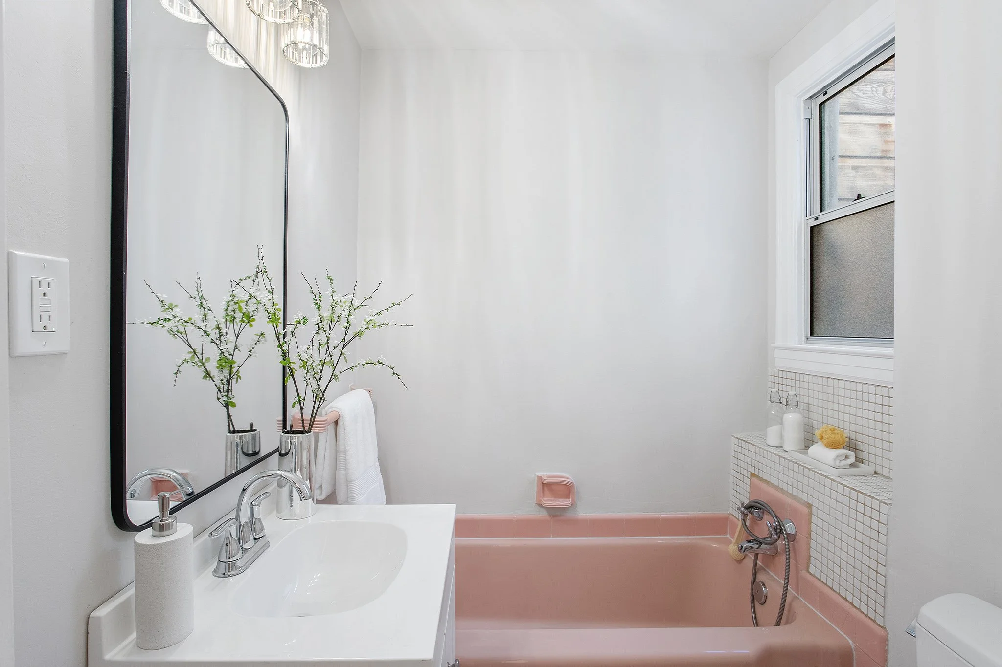 A clean bathroom with a pink bathtub, a window, a mirror with two vases holding branches with small green leaves, a white sink with a soap dispenser, and shelves with white bottles and towels.