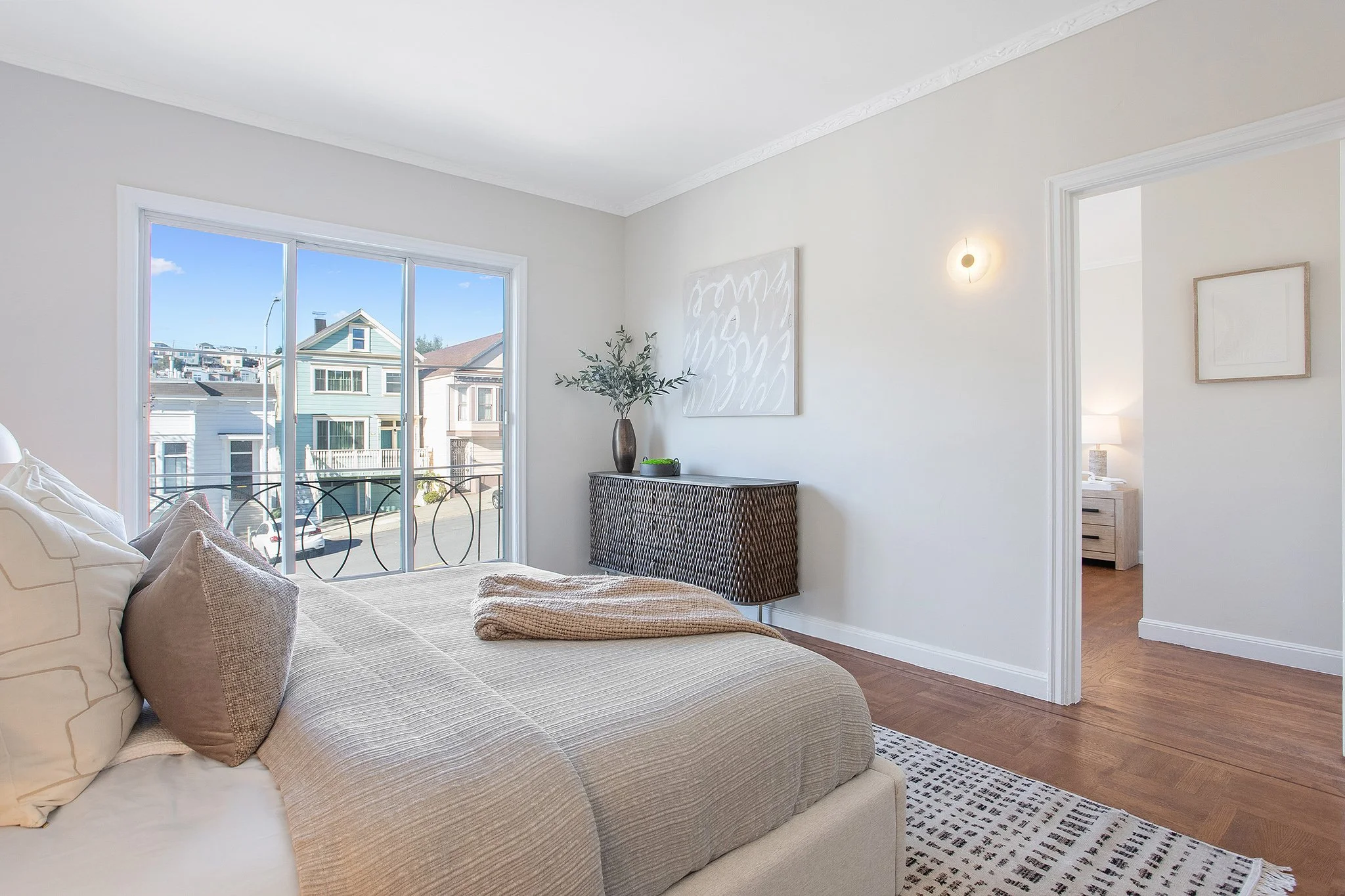 Bright bedroom with a large bed, pillows, a window showing neighboring houses, and minimal decor including a wall art, a side table, and a dresser.