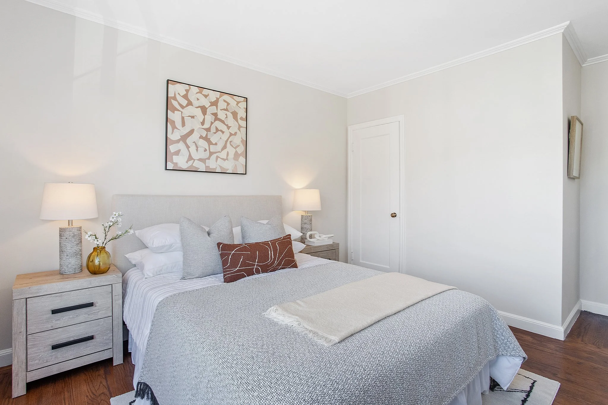 A neatly made bed with a light gray headboard, white bedding, with four pillows and a decorative brown pillow, in a minimalist bedroom with two matching nightstands and lamps, a framed abstract art piece above the bed, and hardwood flooring.