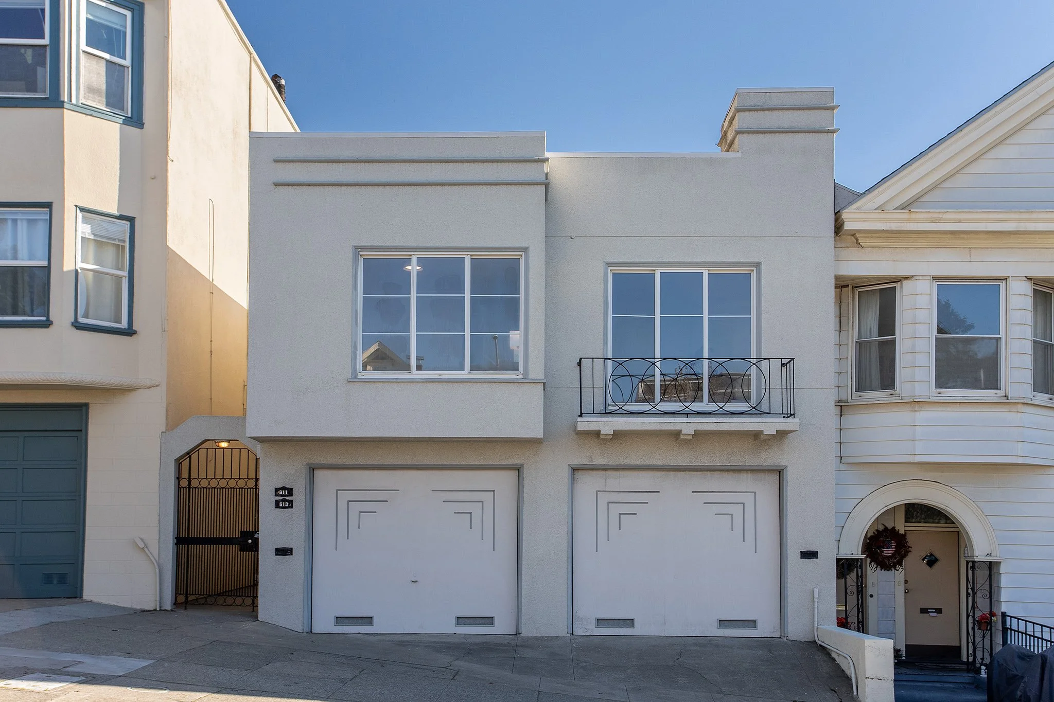 Front view of a modern multi-story white house with two large garage doors, large windows, a small balcony with a black metal railing, and a decorated front door with a holiday wreath.