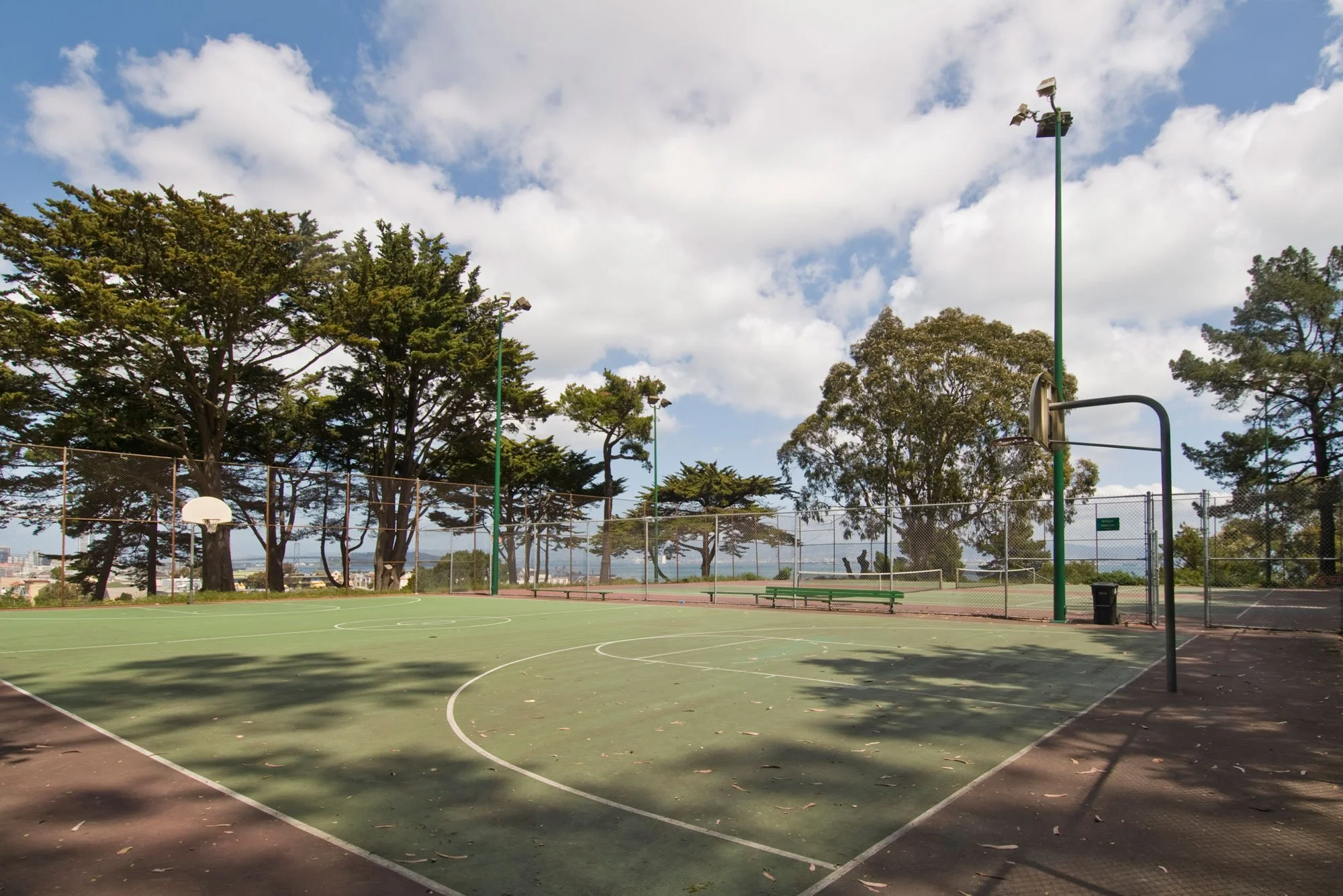 An empty outdoor basketball court surrounded by trees with a city skyline in the distance under a partly cloudy sky.