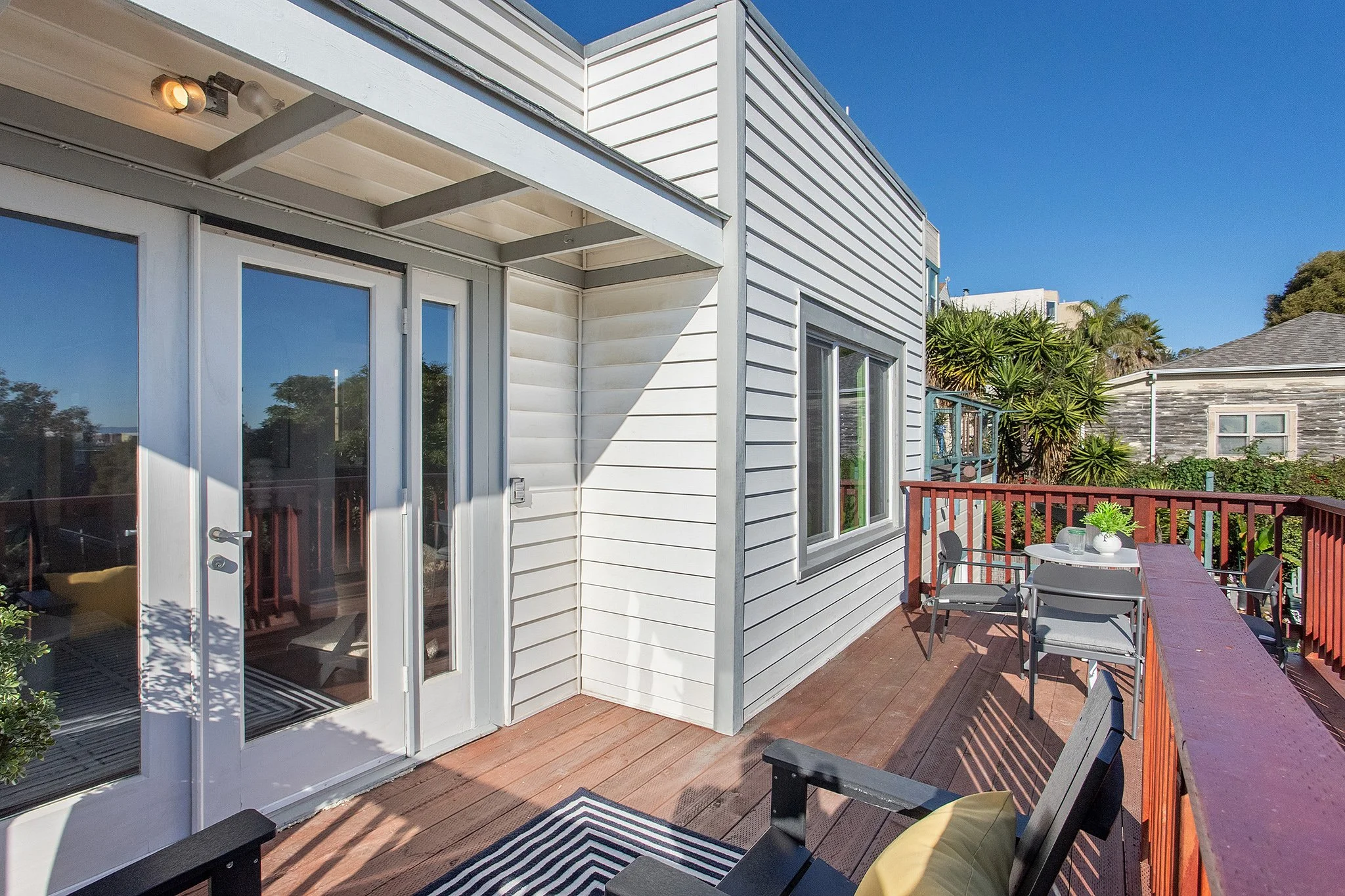 Outdoor balcony with wooden flooring, black chairs, a table with a potted plant, and sliding glass doors on a white house, with neighboring houses and trees in the background under a clear blue sky.