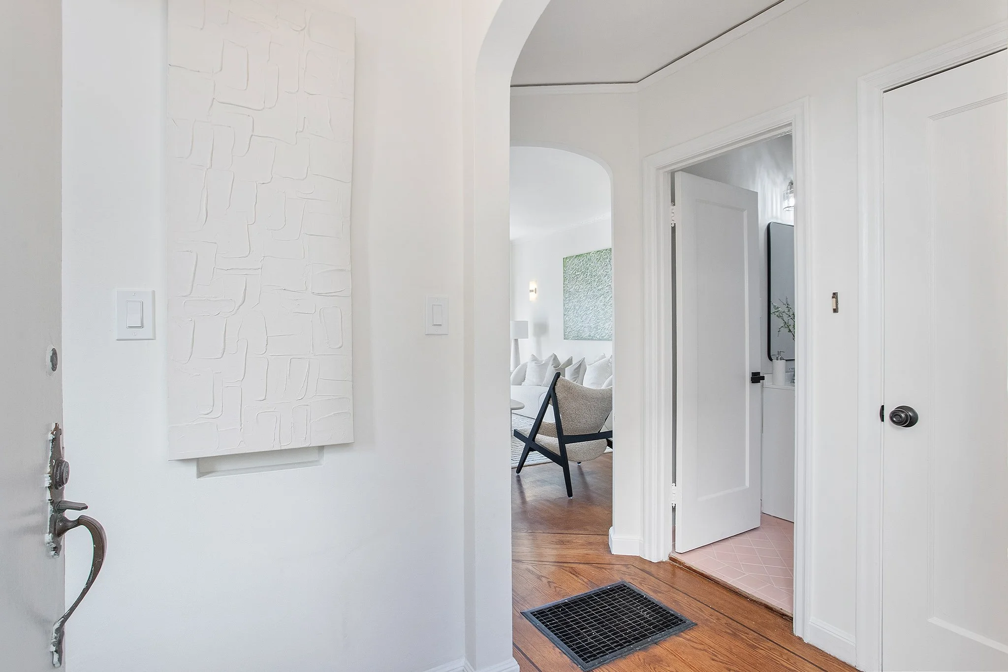 Interior entryway with white walls, a door on the left, and an open doorway leading to a living room with a chair and wall art.