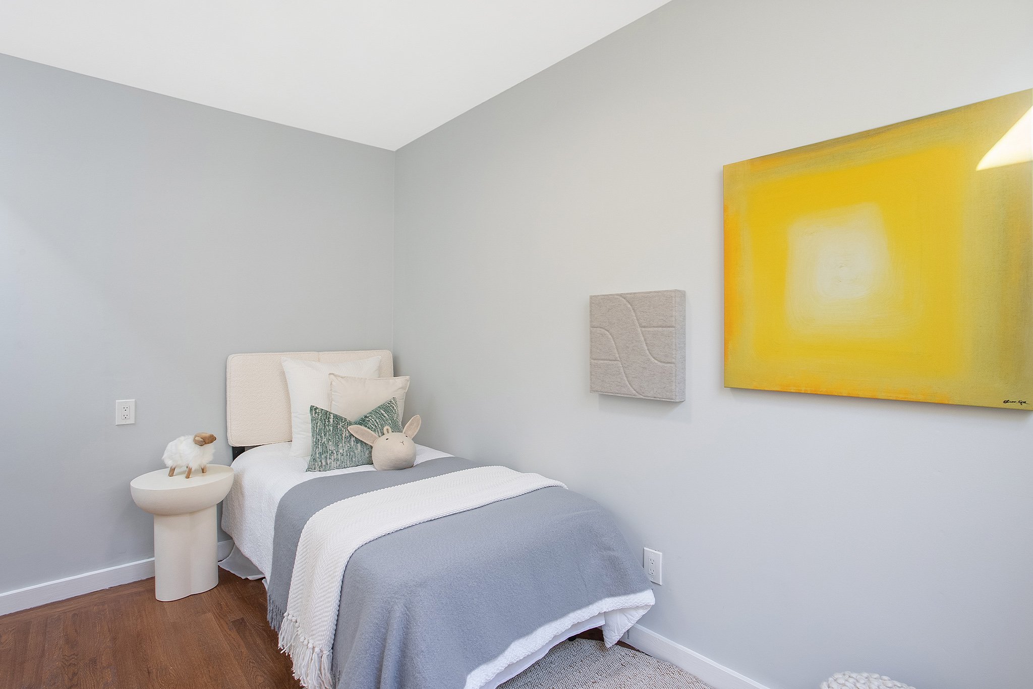 A cozy bedroom with a single bed covered in white and gray bedding, decorated with bunny and sheep plush toys, a white side table, a pastel-colored quilted headboard, wall art including a yellow abstract painting and a grey textured piece, and hardwo