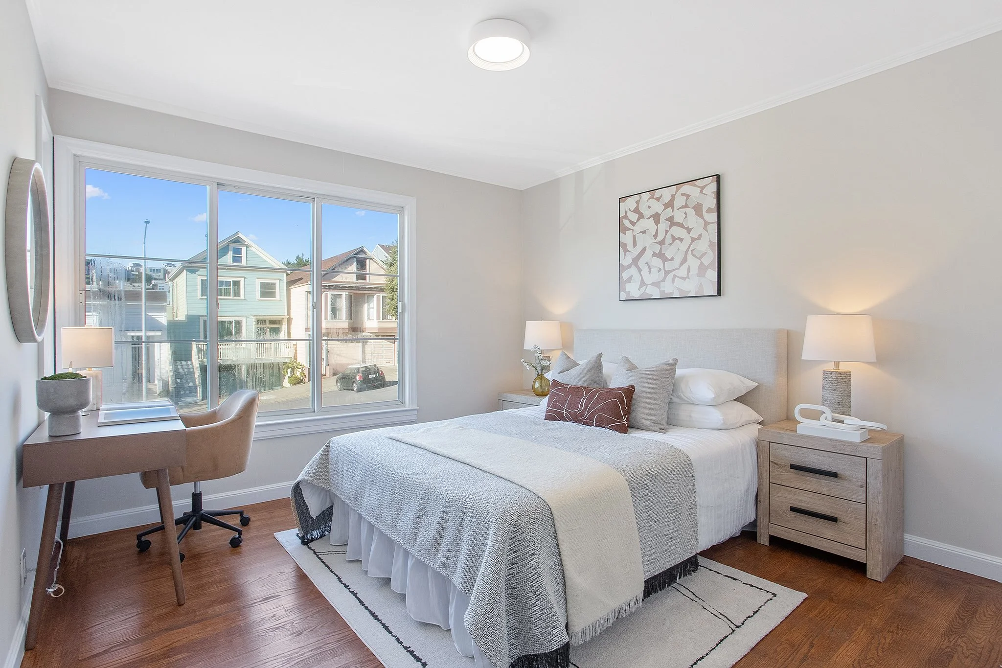 A bright bedroom with a large window showing multi-colored houses outside, a bed with white and beige bedding, and two nightstands with lamps.