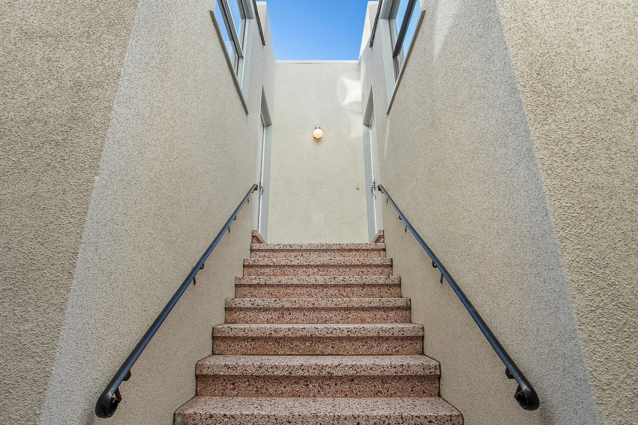 Outdoor staircase with pink granite steps, black handrails, beige stucco walls, leading up to a door with windows, and a blue sky visible at the top