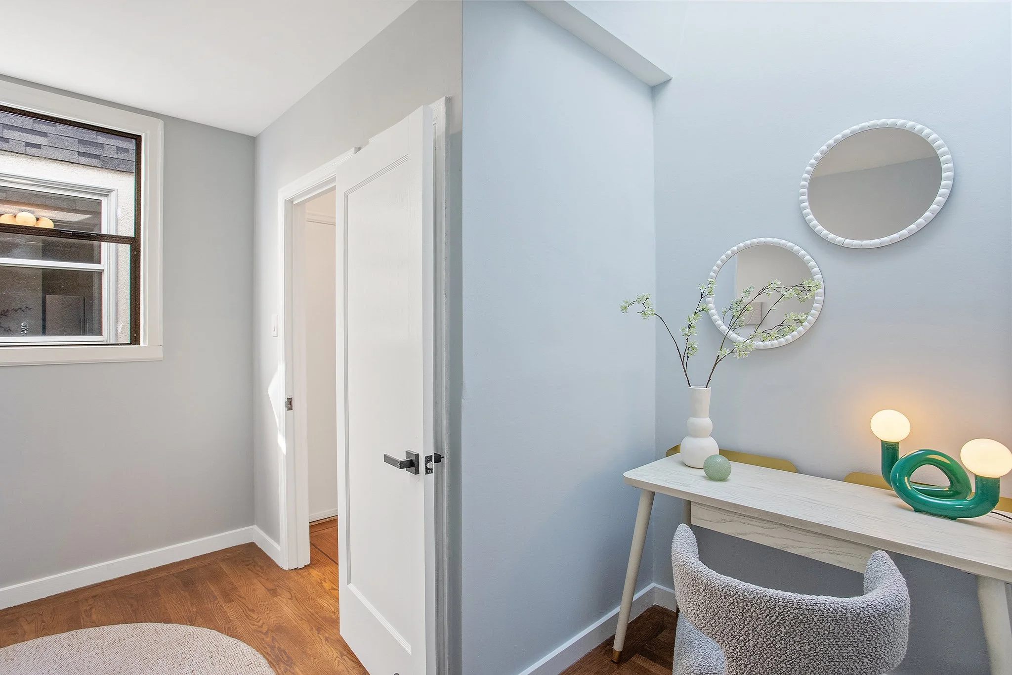 Interior room with a window, white door, light blue walls, two round white framed mirrors, a white marble desk with decorative objects, a chair, and a vase with flowering branches.
