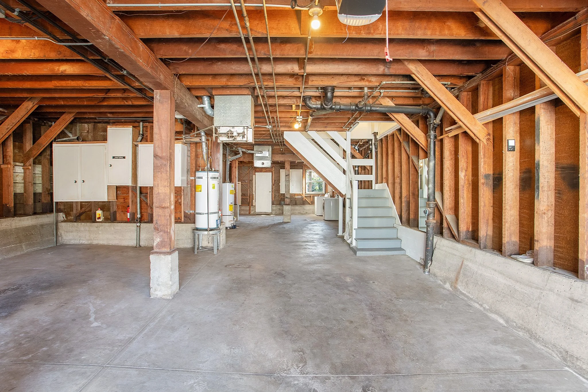 Empty unfinished garage with exposed wooden beams, concrete floor, water heaters, and utility pipes.