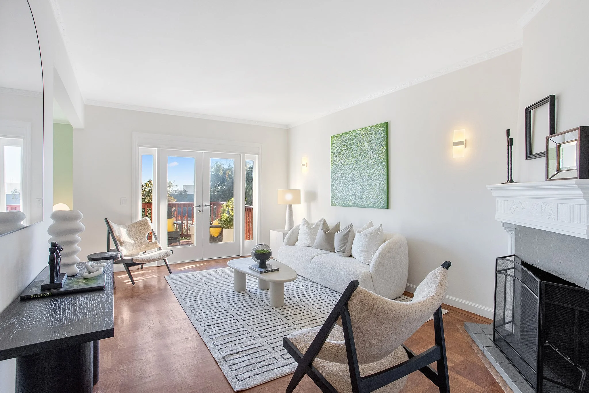 Bright living room with white walls, wooden floor, glass double doors leading to a balcony, and modern furniture including a white sofa, beige armchairs, a black coffee table, and a black console table. Decor includes artwork, sculptures, and a firep