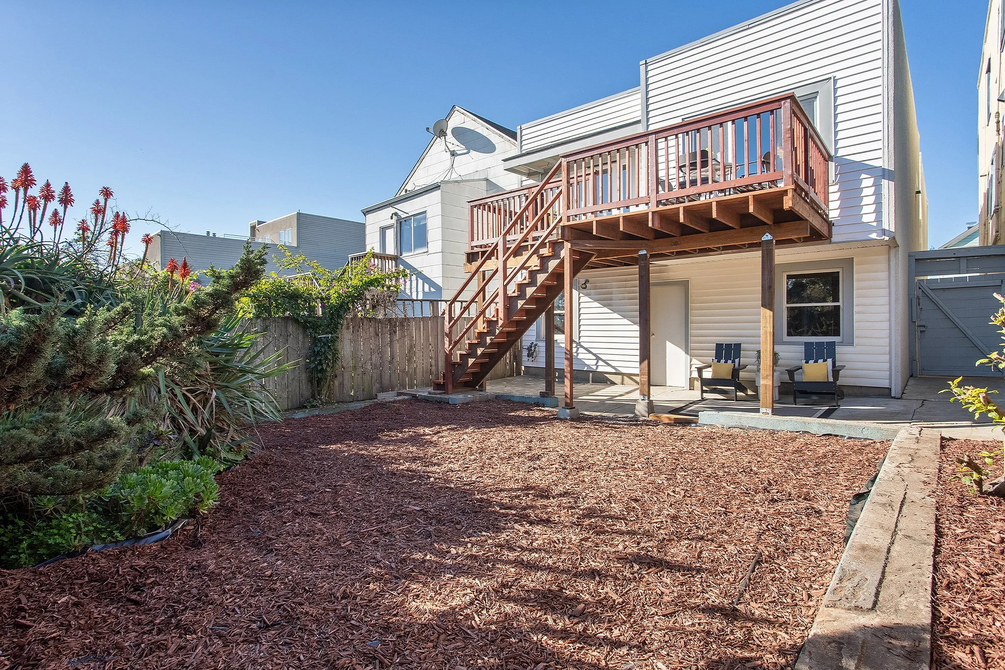 Backyard with a patio, two chairs, an upper wooden deck with stairs, beige house with white siding, and a garden with plants and mulch.