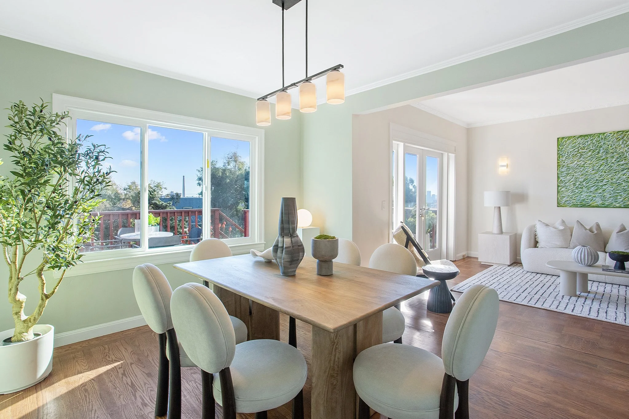 Interior of a bright, modern living and dining room with large windows, a wooden dining table with six white upholstered chairs, a white sofa with white pillows, a striped rug, and decorative vases and sculptures.