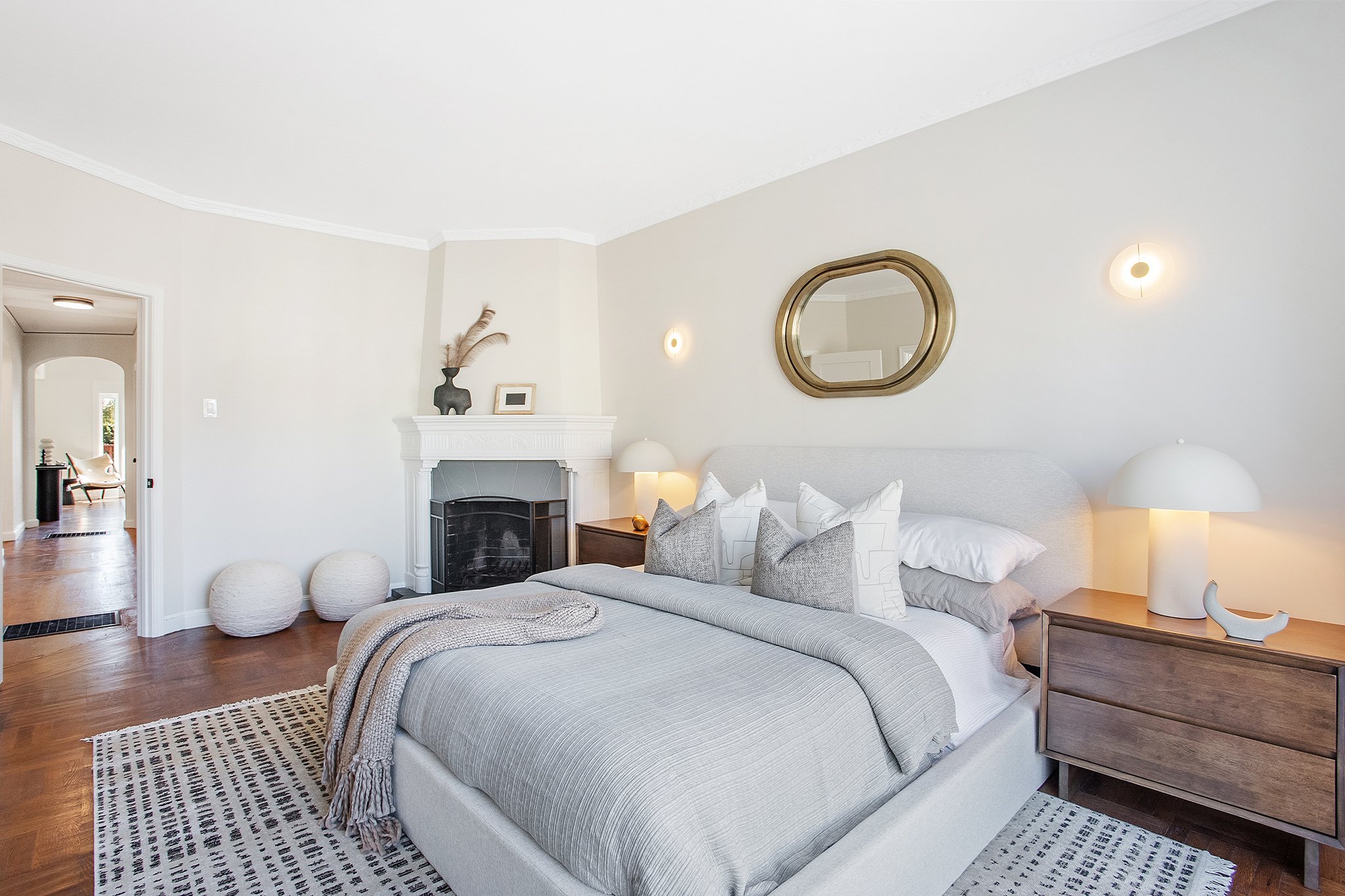 A modern bedroom with a large bed, white bedding, decorative pillows, two wooden nightstands with white lamps, a mirror above the bed, a fireplace with decorations, and a doorway leading to another room with a chair.
