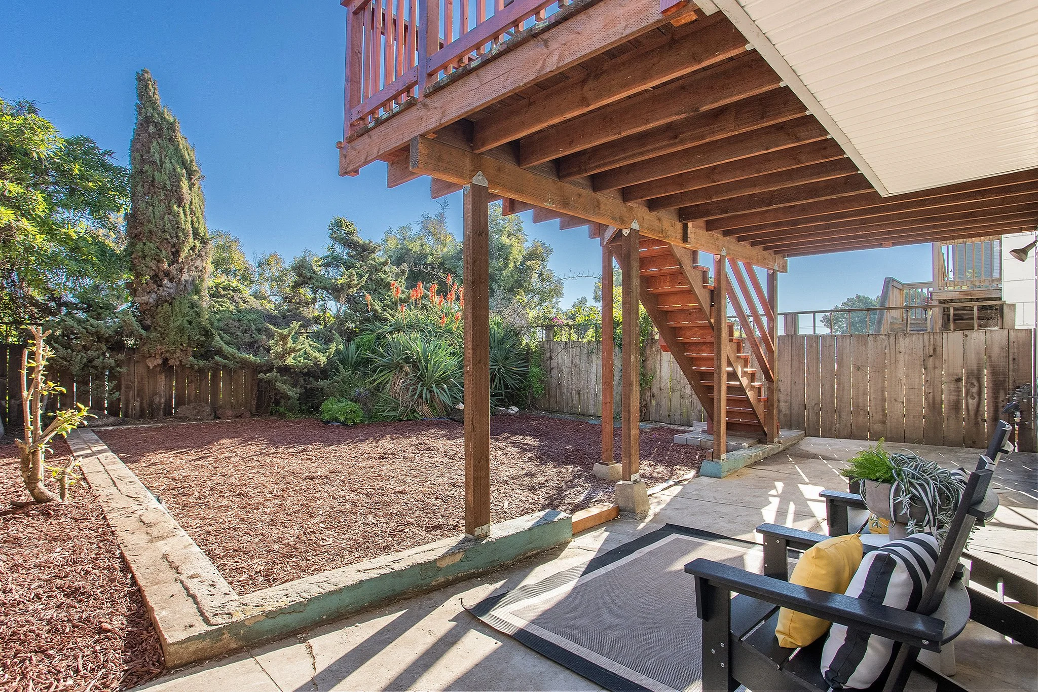 Backyard patio with a wooden staircase, outdoor seating with striped cushions and yellow pillows, fenced yard with trees and bushes, clear blue sky.